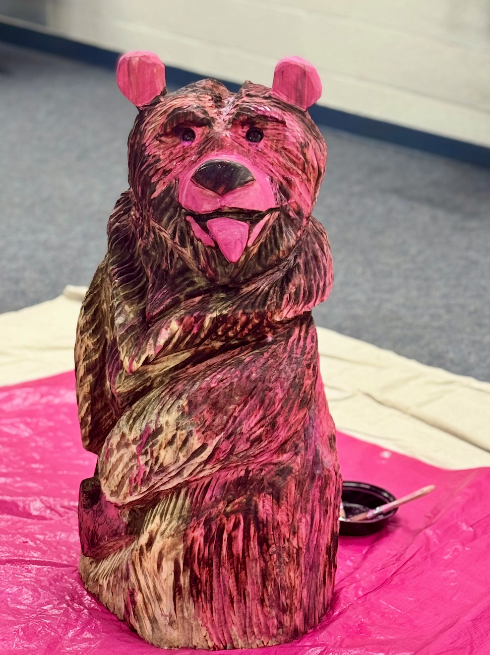 Wooden bear sculpture painted with pink and brown colors, with a pink tongue, placed on a pink cloth with a paintbrush and black paint on a small dish nearby. (Painting Pynko Pynk - currently in progress)