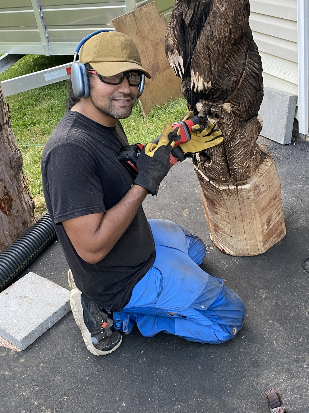 Sawtistic is kneeling on one knee, wearing a cap, safety glasses, headphones, gloves, black t-shirt, and blue work pants, is using a power tool to carve details into a large wooden sculpture of an animal, likely a bear, outdoors near a house.