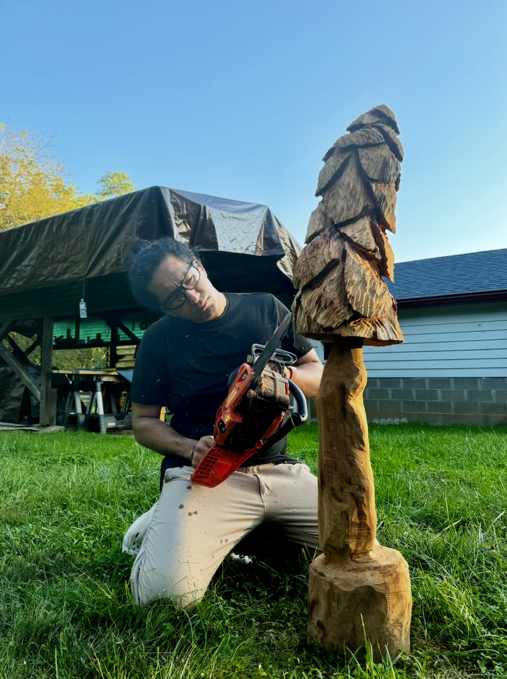 Sawtistic is kneeling on grass, using a chainsaw on a carved wooden sculpture of a tree, outdoor setting with a house, trees, and blue sky in the background.