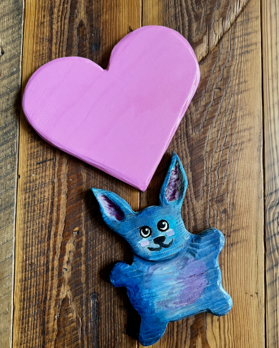 Hand-painted wooden heart painted pink and a colorful painted rabbit figurine on a wooden surface.