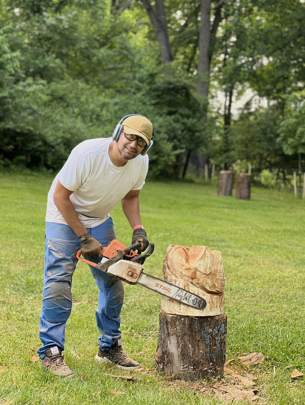 Sawtistic is wearing a beige cap, safety glasses, ear protection, a white T-shirt, and blue pants and is cutting a piece of wood with an orange chainsaw in a grassy outdoor area with trees in the background.