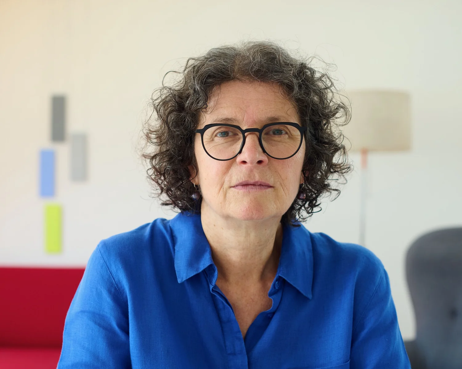 Nahes Porträt von Elisabeth Heindl im blauen Hemd, mit farbigen Arbeiten und einem roten Sofa im Hintergrund.