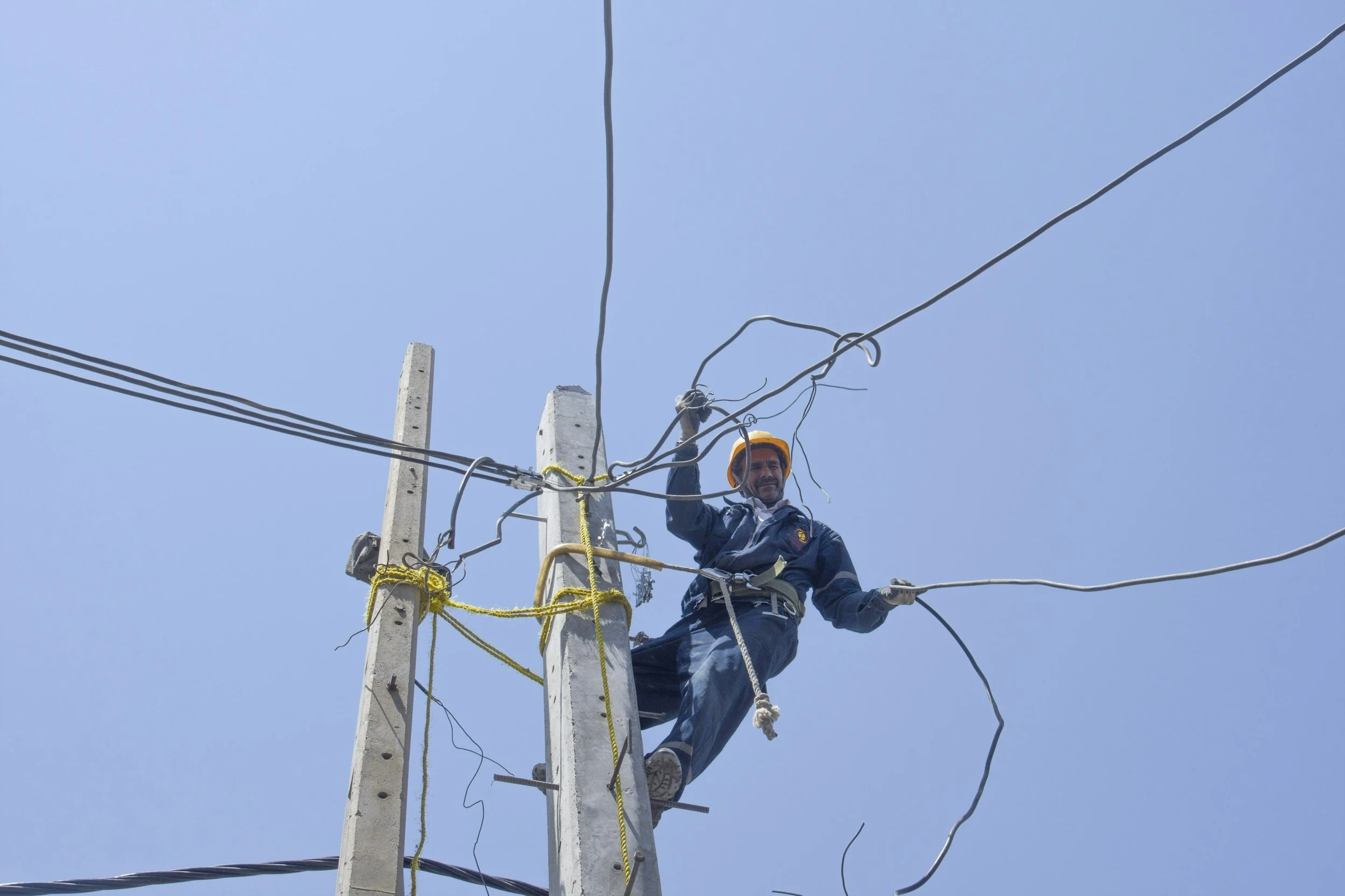 A utility worker wearing a yellow safety helmet and blue uniform working on electrical wires atop a utility pole against a clear blue sky.
