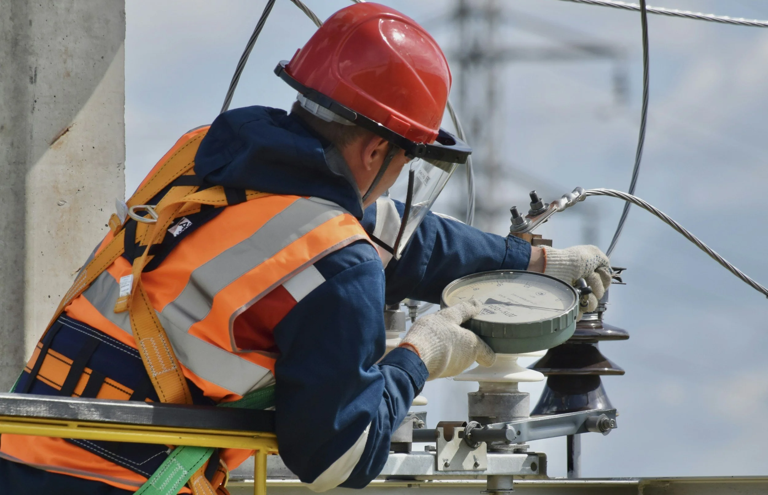An electrician wearing a red safety helmet, orange high-visibility vest, and gloves working on electrical equipment outdoors.