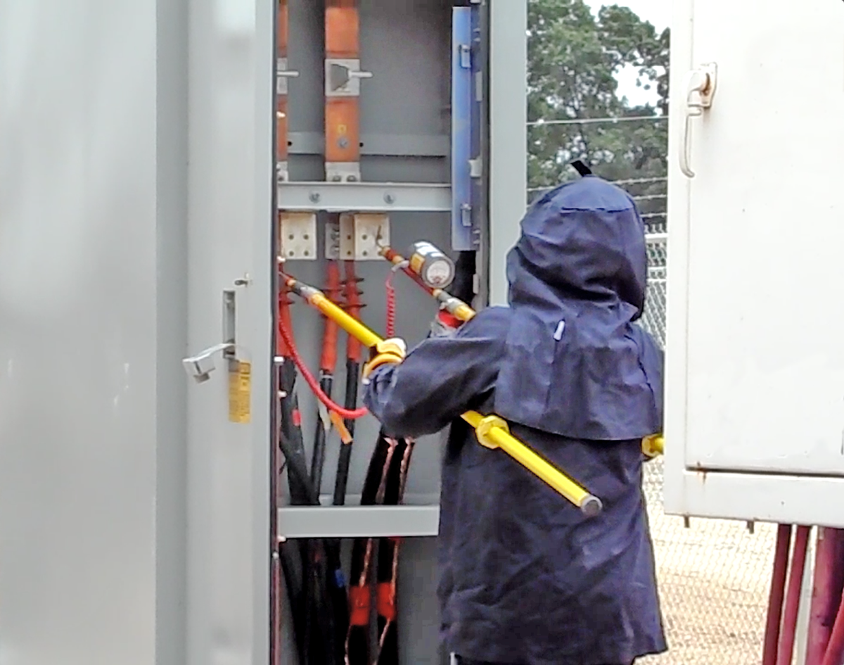A technician wearing a blue hooded rain jacket working on electrical equipment inside an open electrical box outdoors.