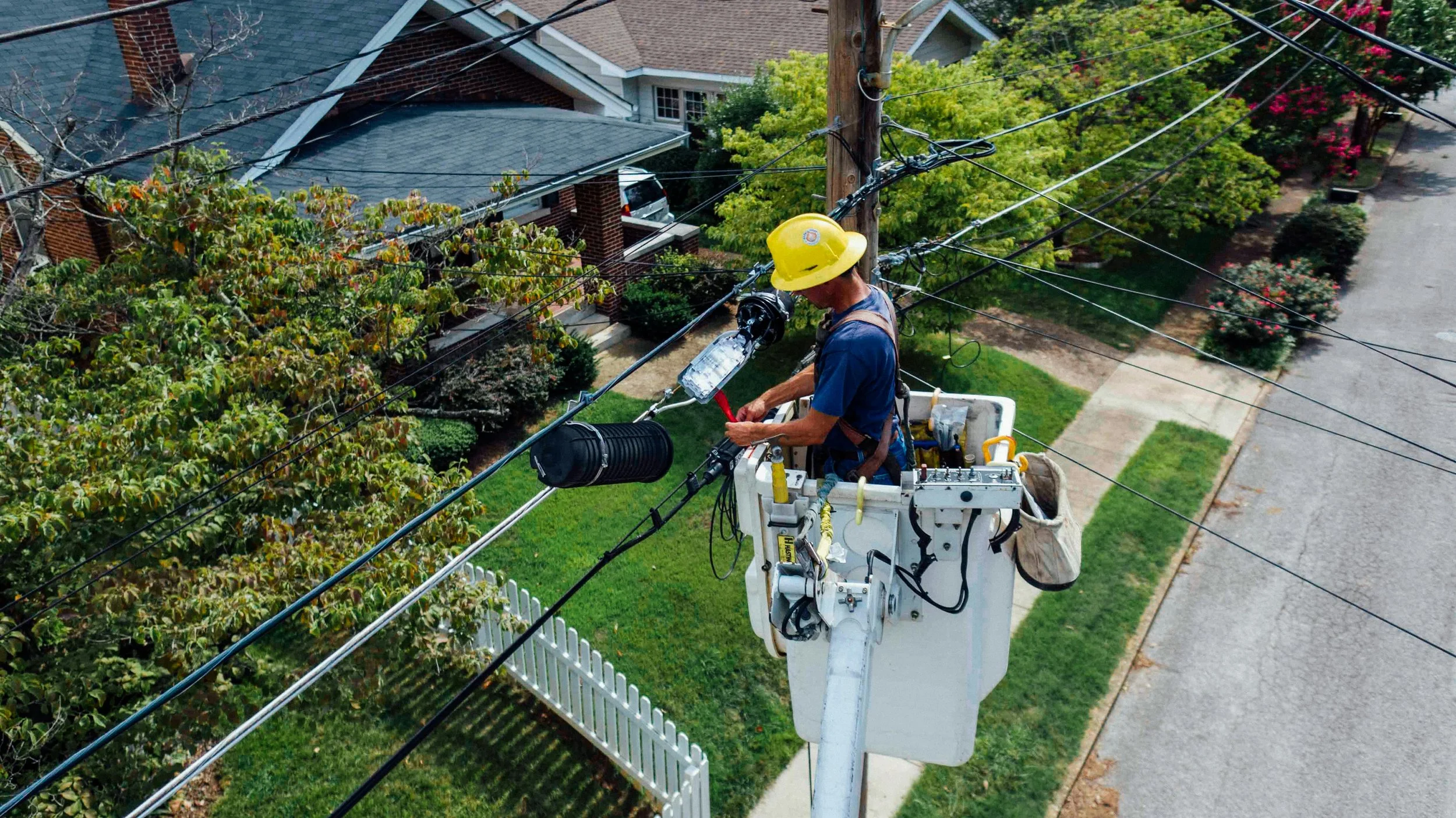 A utility worker wearing a yellow hard hat and blue shirt working on power lines from a lift bucket in a residential neighborhood