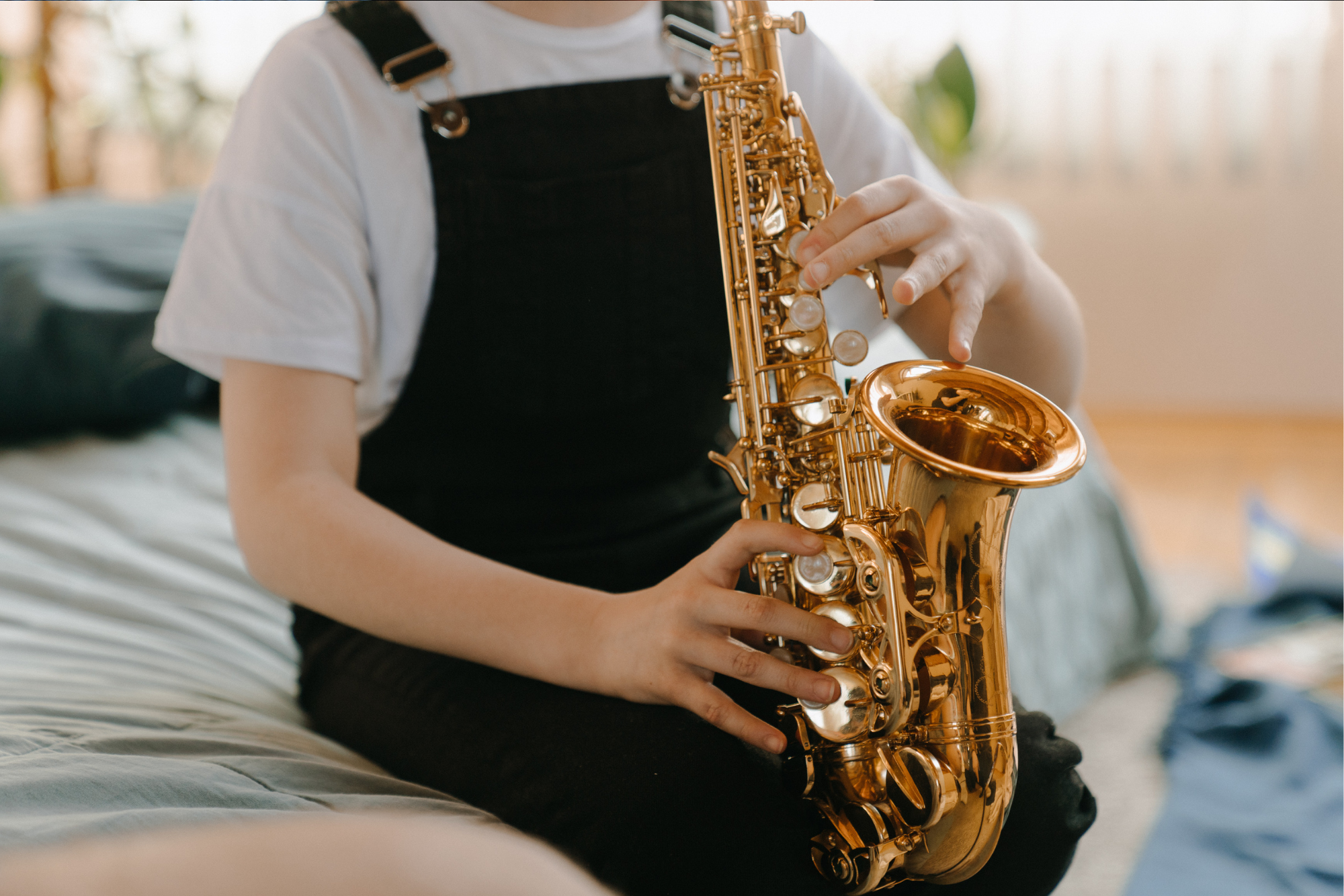 Child playing gold-colored saxophone while sitting on a bed, with a blurred background of a room with natural light.