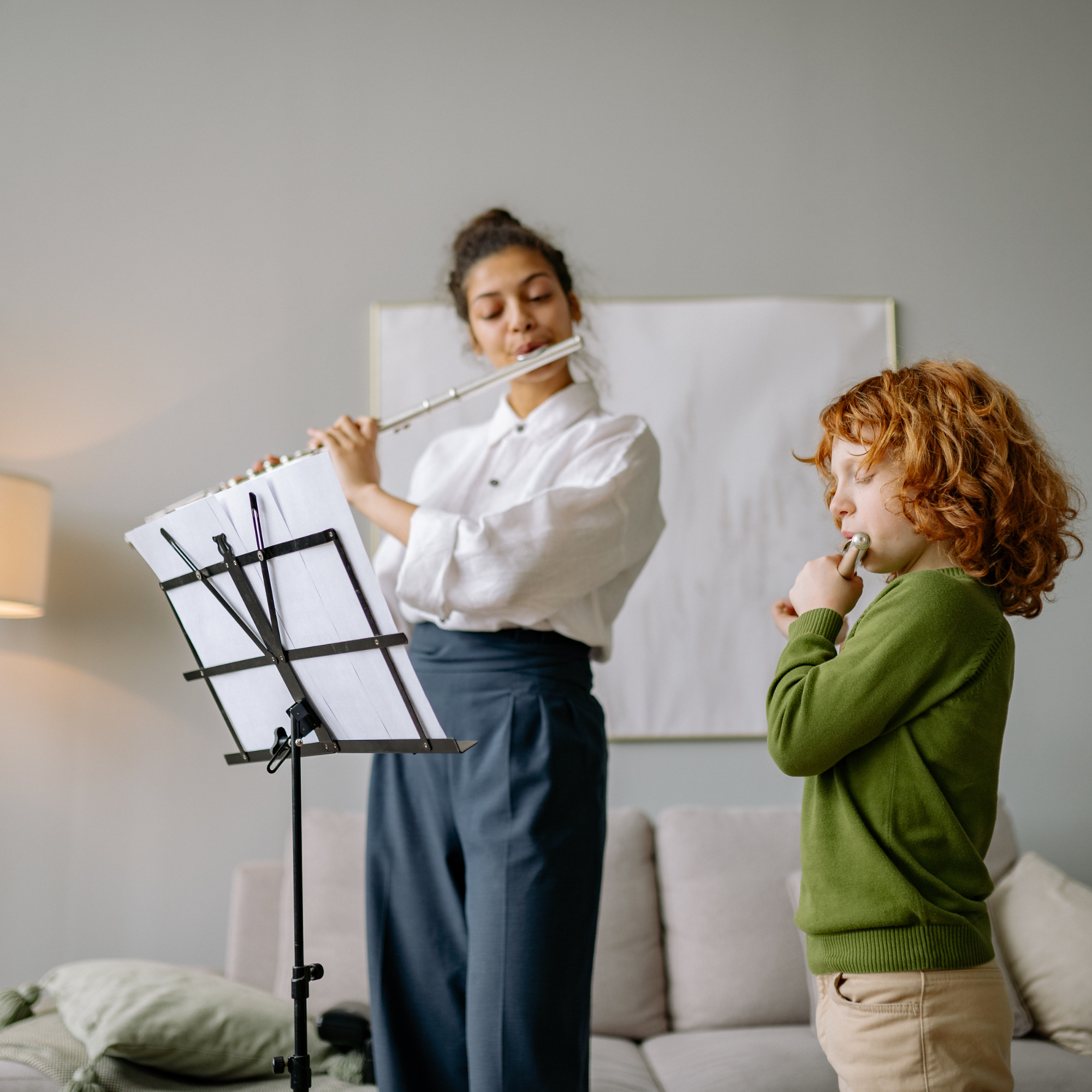 A woman and a young boy playing flutes in a living room with a music stand, a sofa, and a lamp.