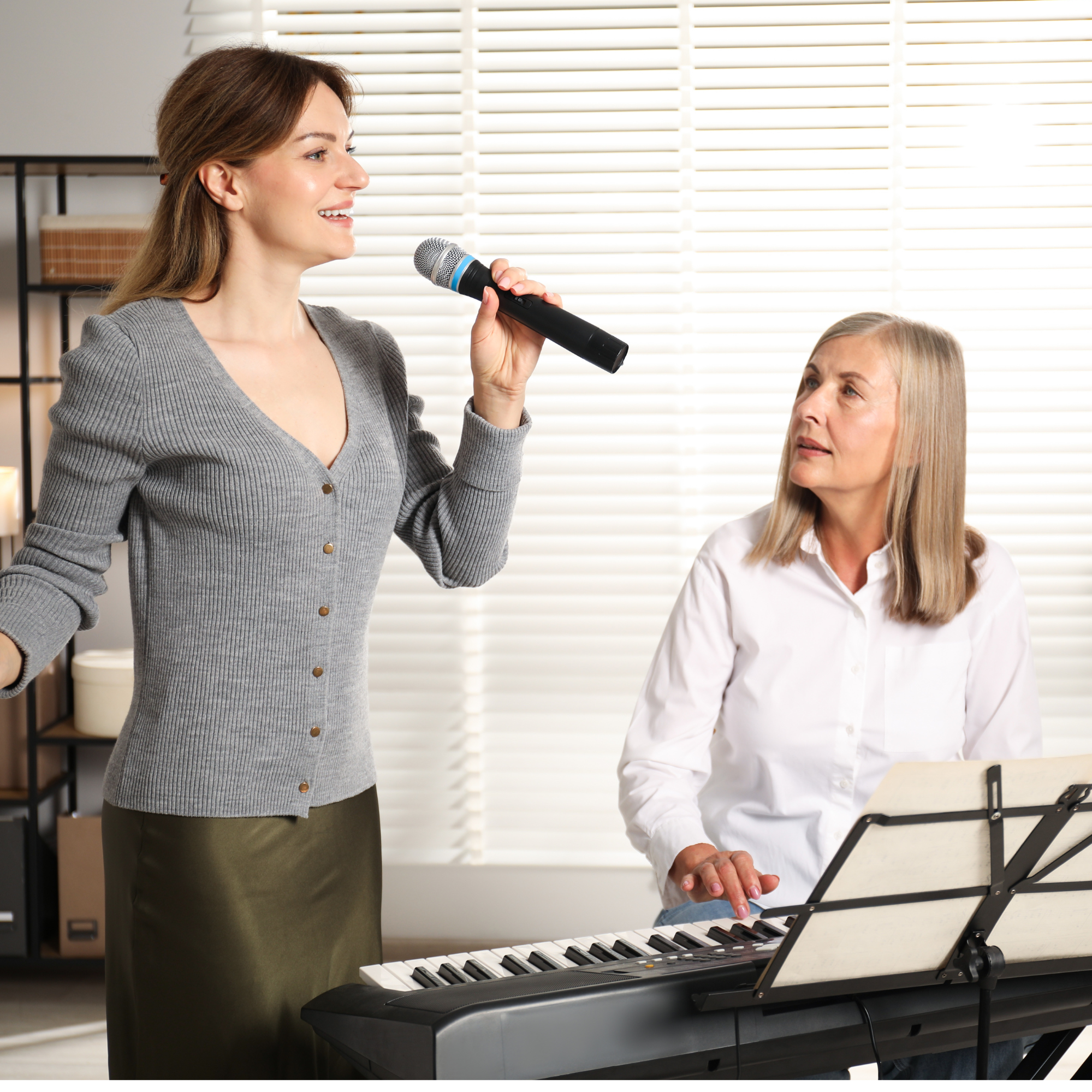 A woman with a gray cardigan and wearing lipstick holds a microphone and sings or speaks, while another woman with blonde hair wearing a white shirt plays a keyboard and looks at her. They are in a room with white blinds and some shelves.