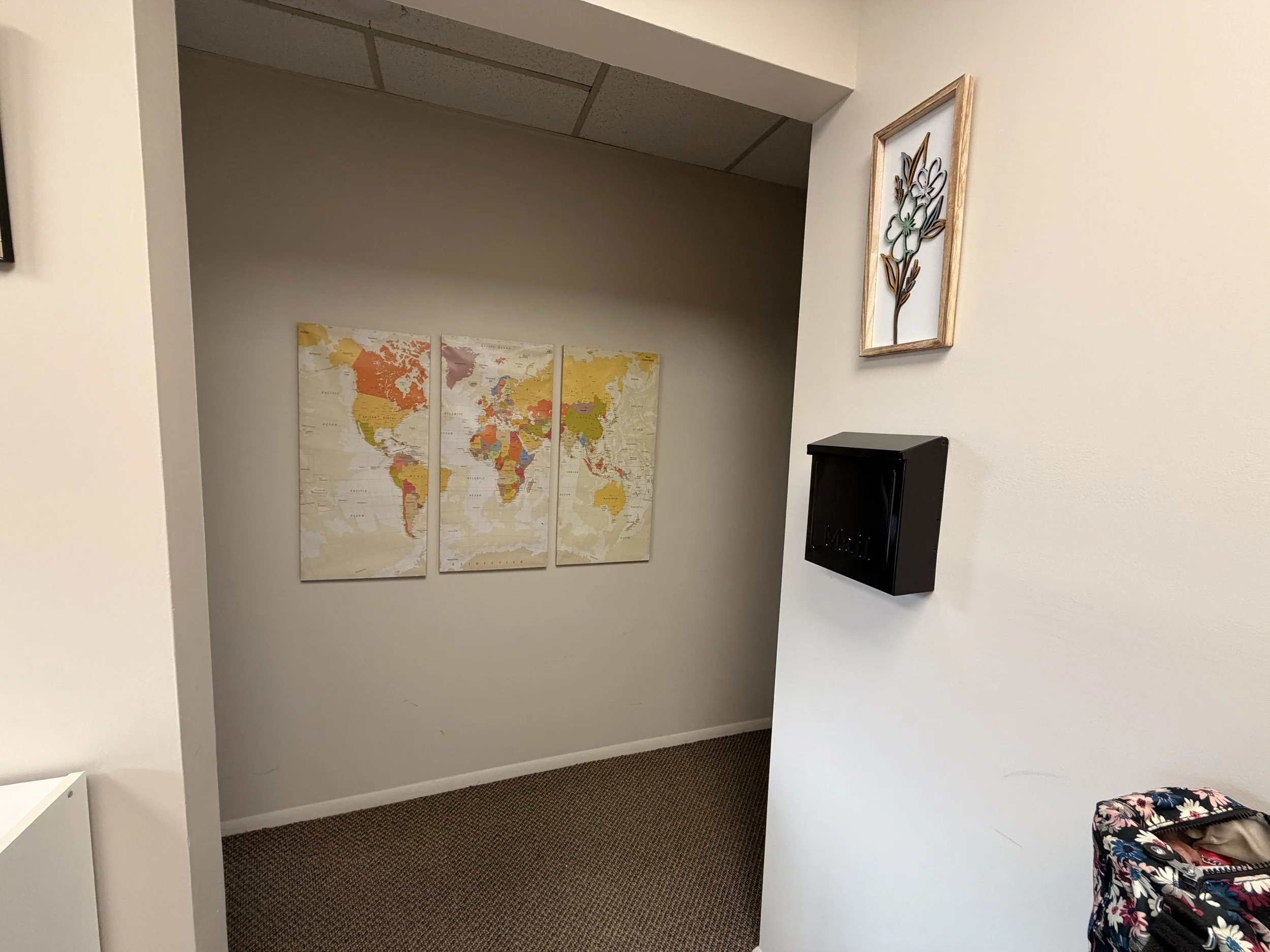 A beige-colored room with a three-panel world map on the back wall, a small framed floral artwork on the right wall, a black mailbox, and part of a floral-patterned bag in the bottom right corner.