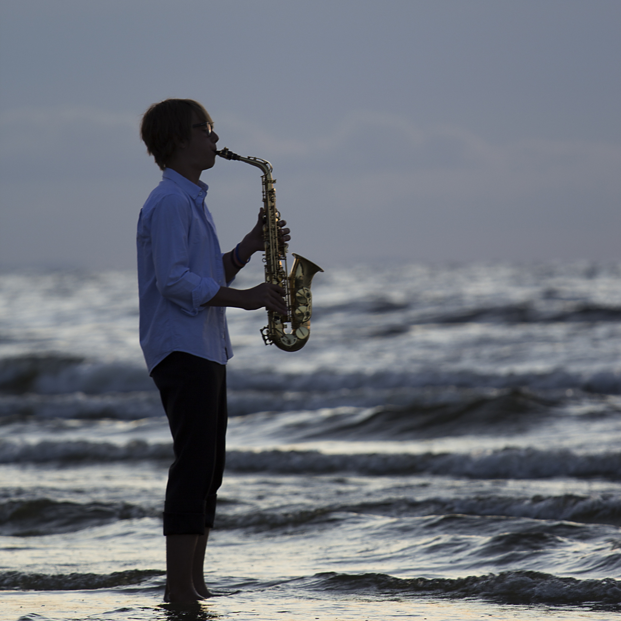 Person standing in the ocean at sunset, playing a saxophone.