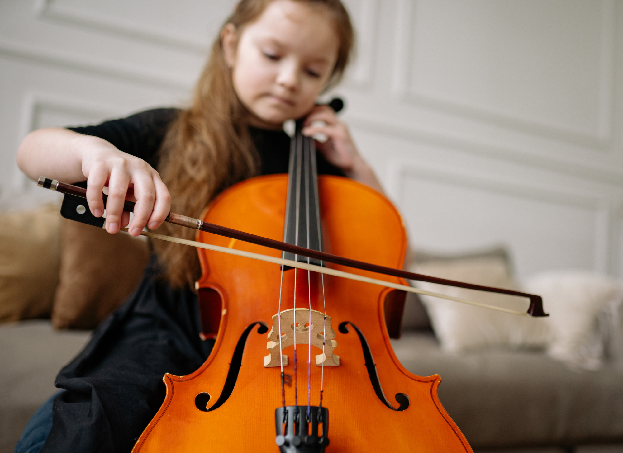 A young girl with long, reddish hair playing a bright orange cello indoors.