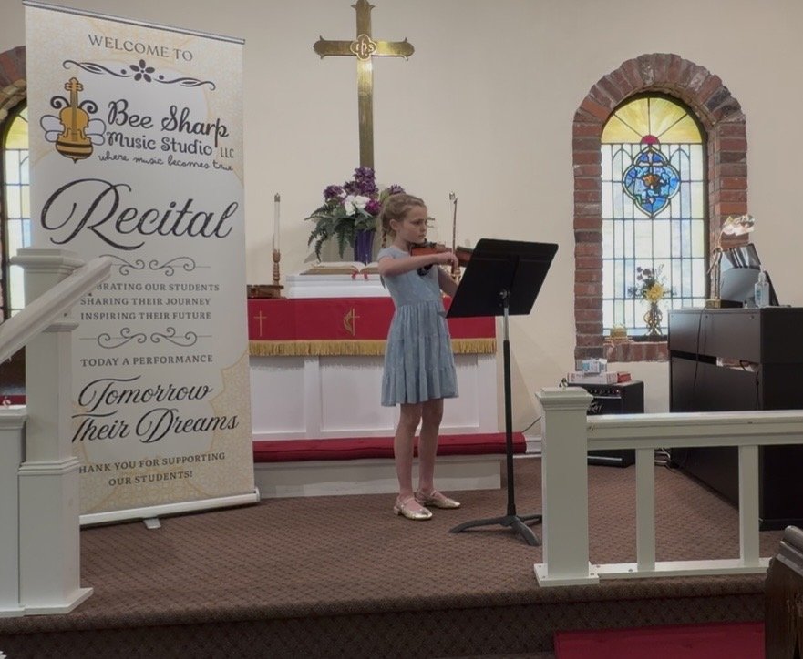 A young girl playing the violin during a recital inside a church or chapel, with a stained glass window and a cross on the wall behind her, and a large banner beside her that reads 'Bee Sharp Music Studio LLC Recital'.