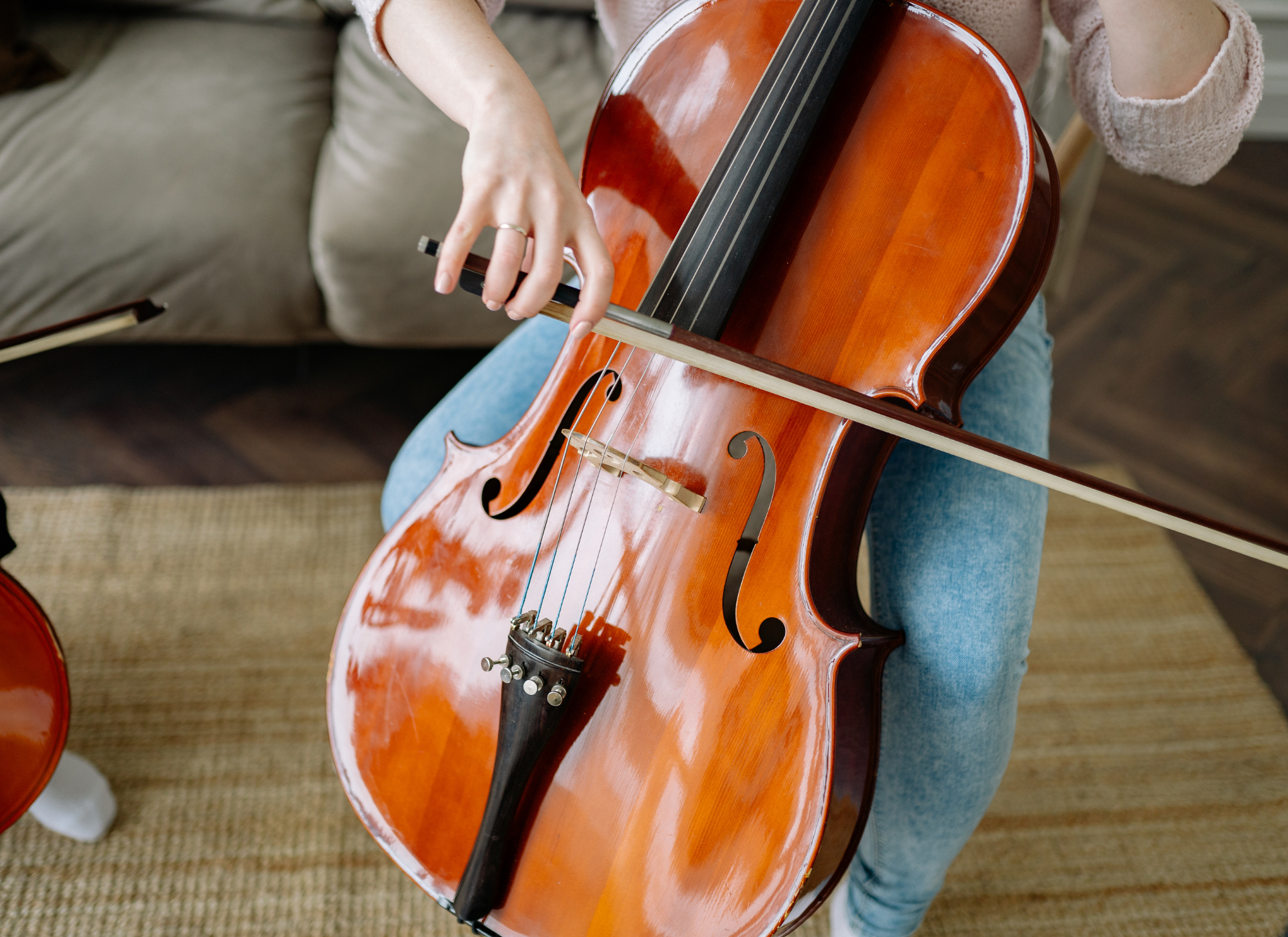 Person tuning a cello indoors, sitting on a chair, with a sofa and a woven rug visible in the background.