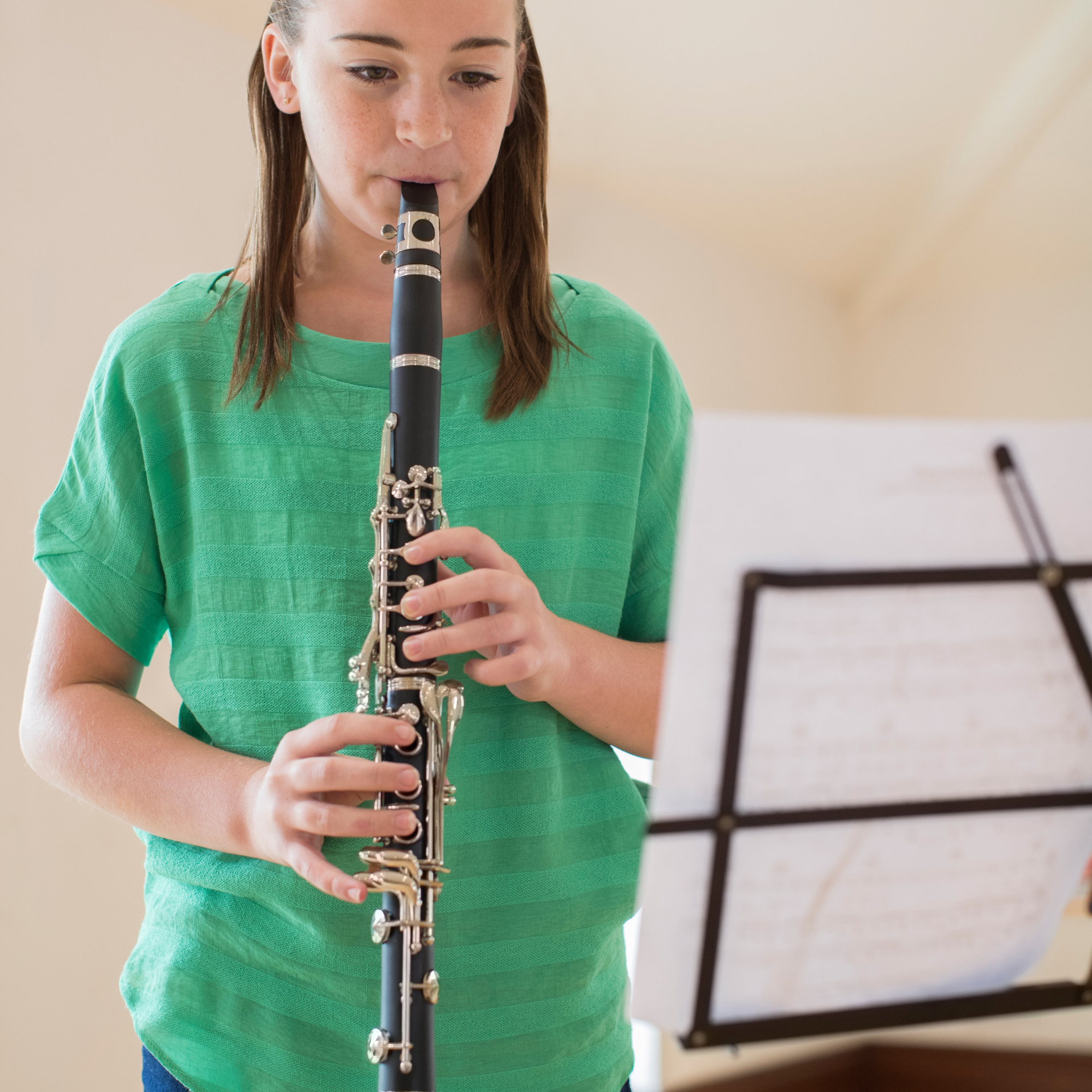 A girl in a green shirt playing a black and silver clarinet with a music stand in front of her.
