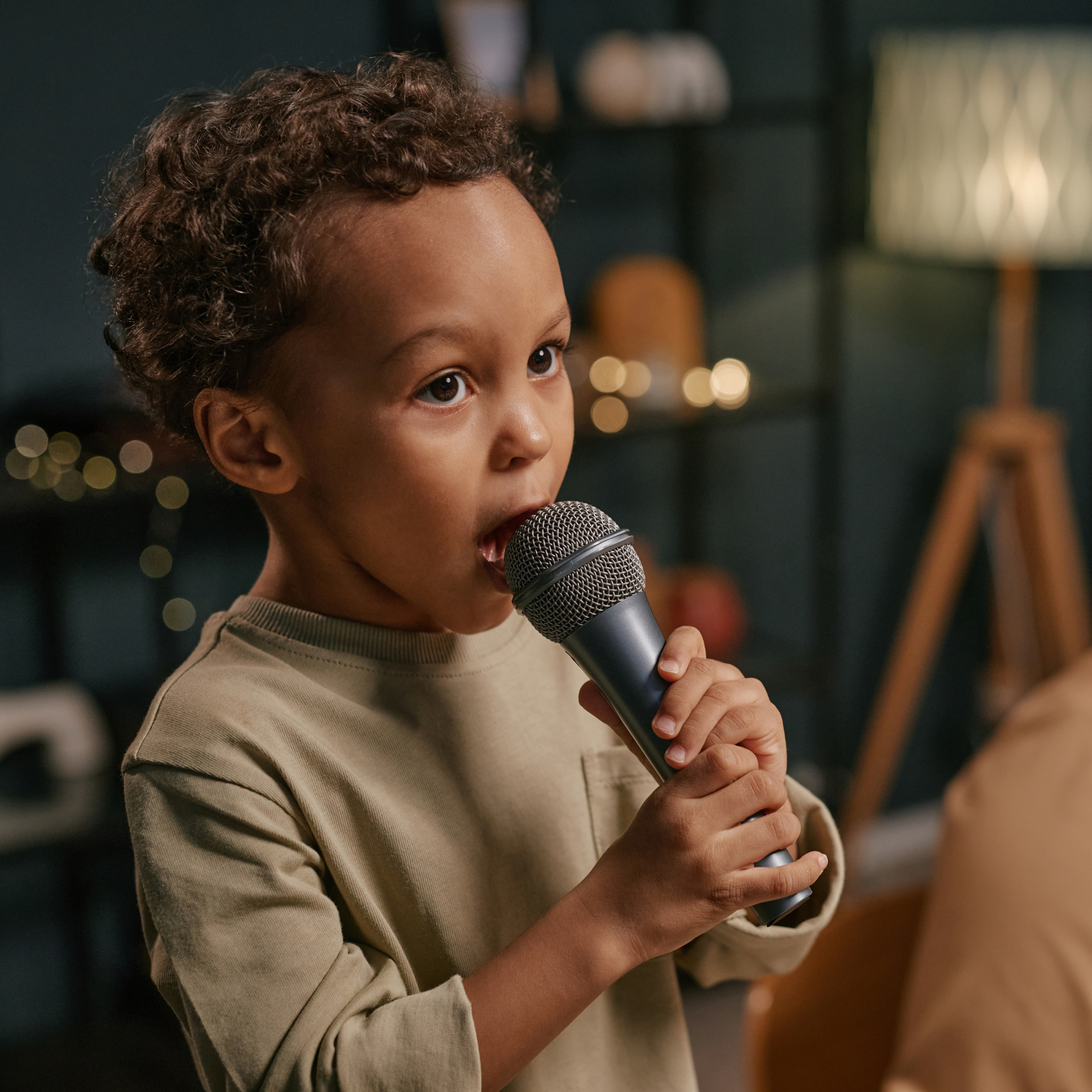 A young boy with curly hair holding a microphone close to his mouth, likely singing or speaking in a cozy indoor setting.