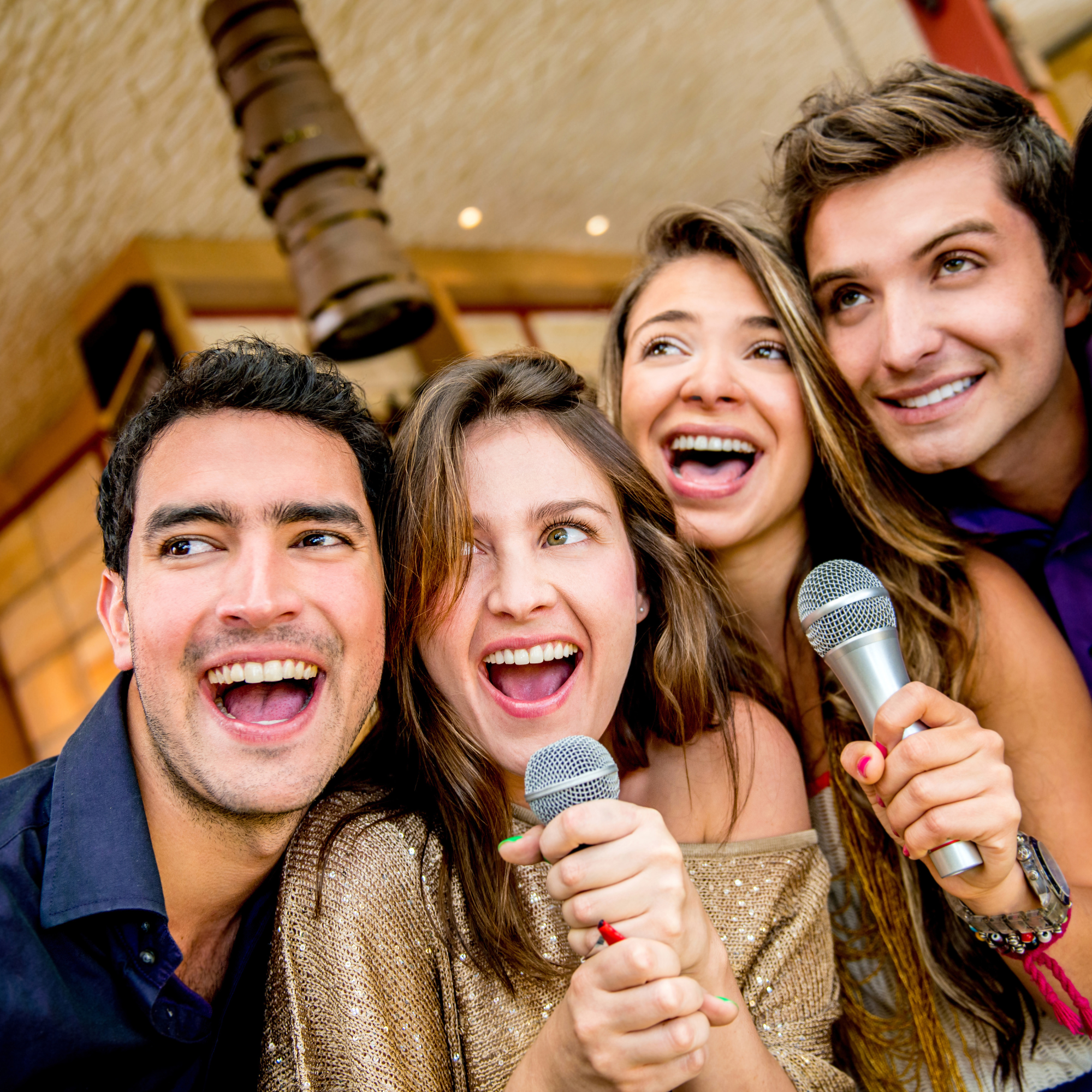 Four young adults smiling and holding microphones at a lively gathering or karaoke event in a warm, cozy indoor setting.