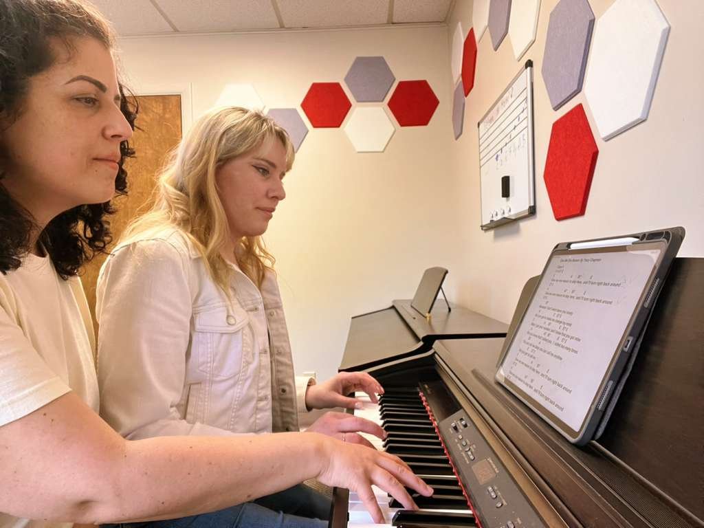 Two young women practicing piano together in a music room with hexagon wall decorations and a whiteboard.