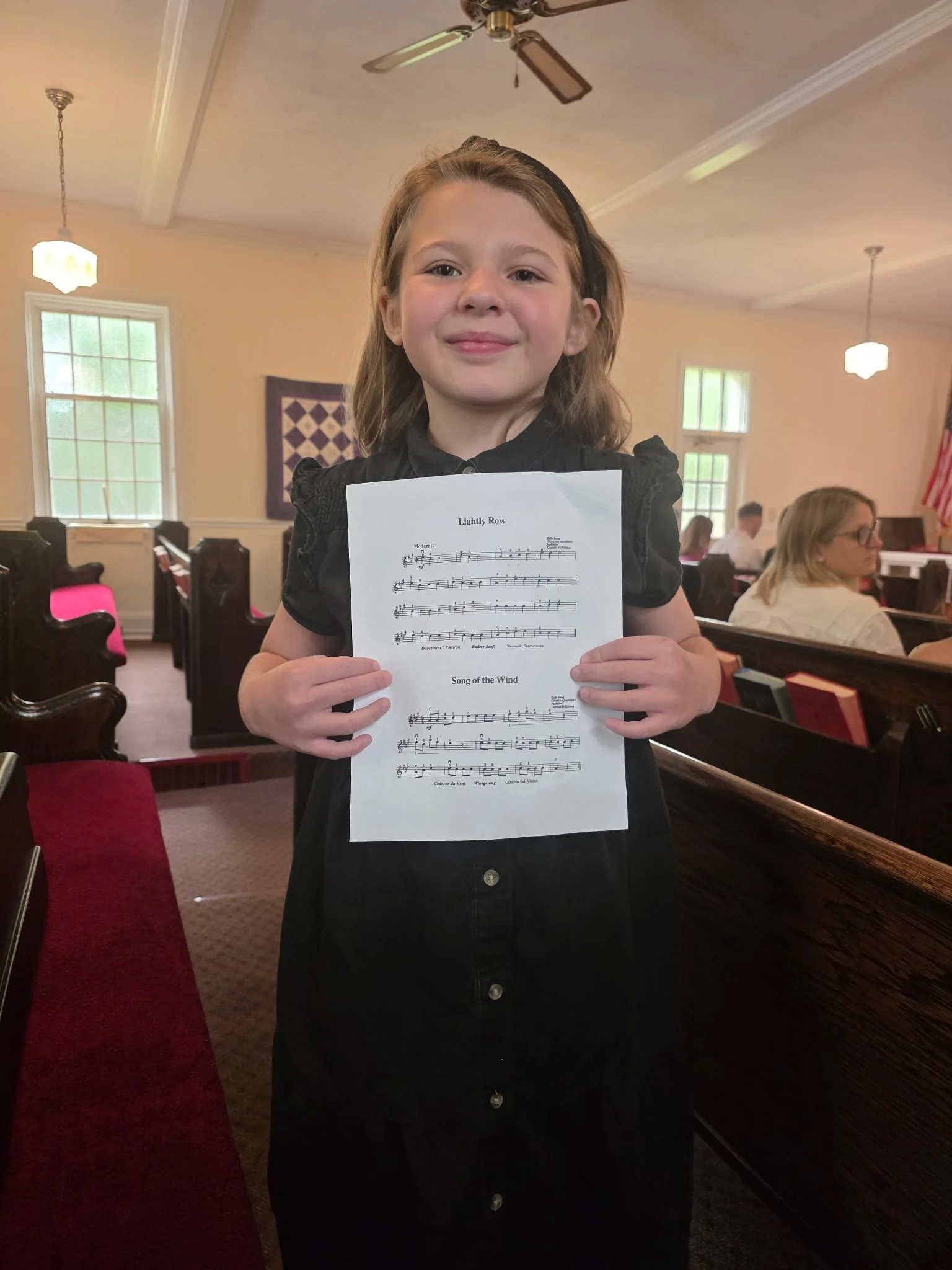 A young girl with light brown hair wearing a black dress holding a sheet of music titled "Song of the Wind" inside a church or chapel with wooden pews, windows, and a few adults seated in the background.