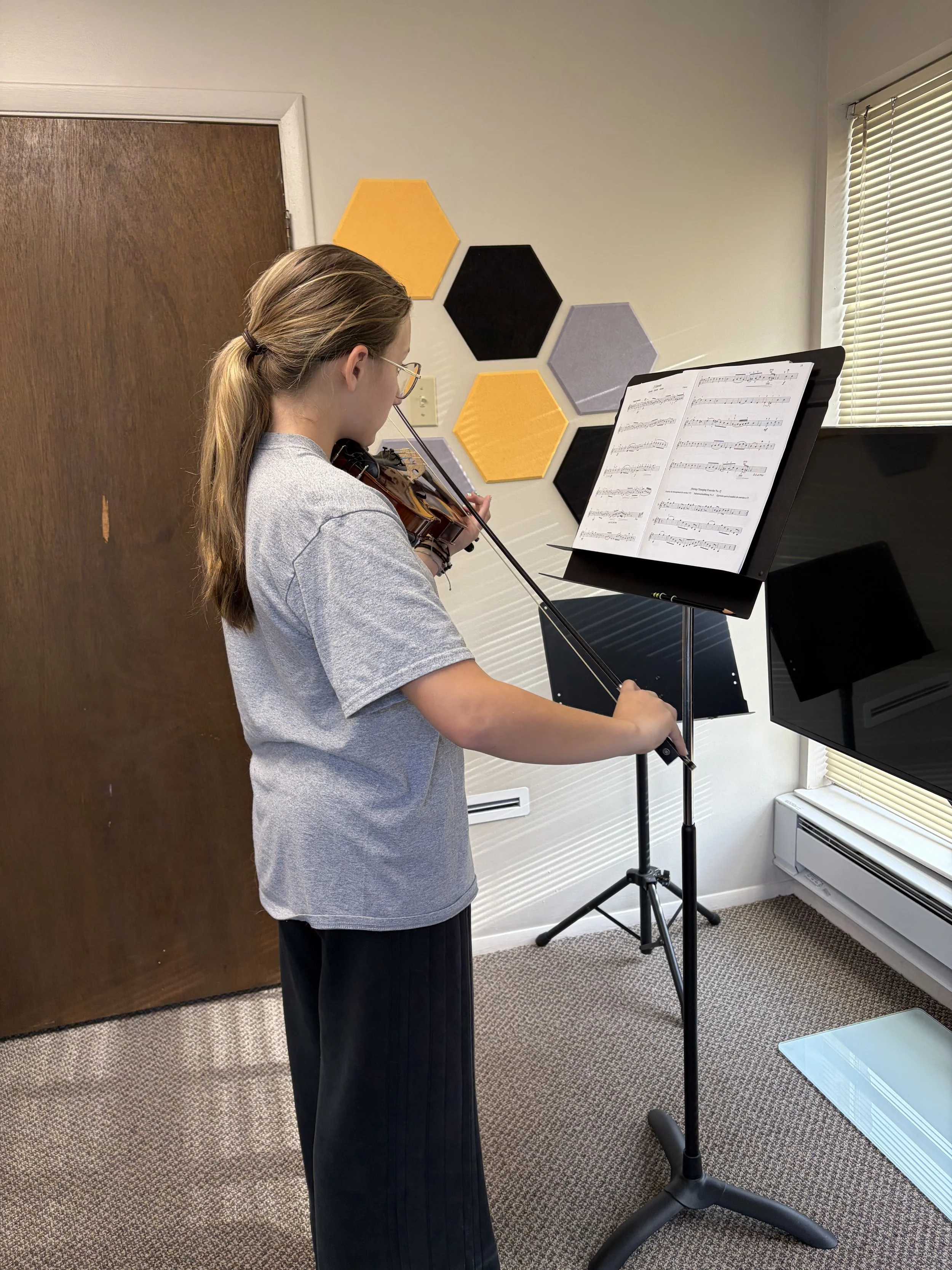 Young woman with brown hair in a ponytail, wearing glasses, playing the violin while reading sheet music on a music stand in a room with hexagonal wall decorations.