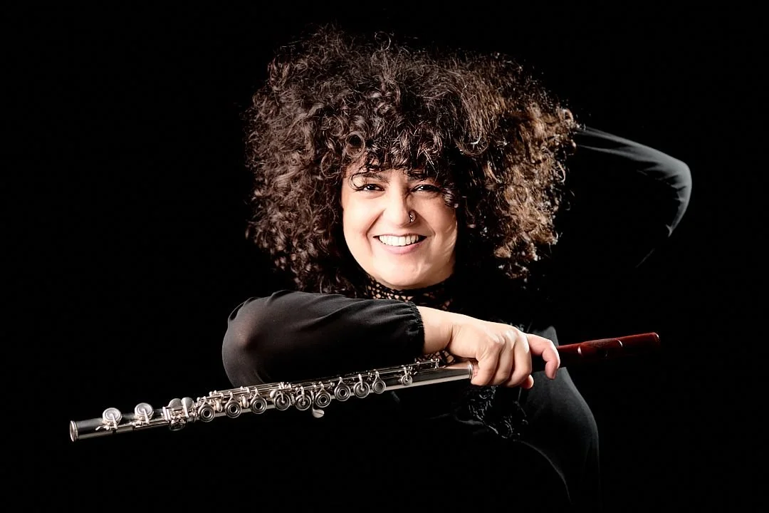 A woman with curly hair smiling and holding a flute against a black background.