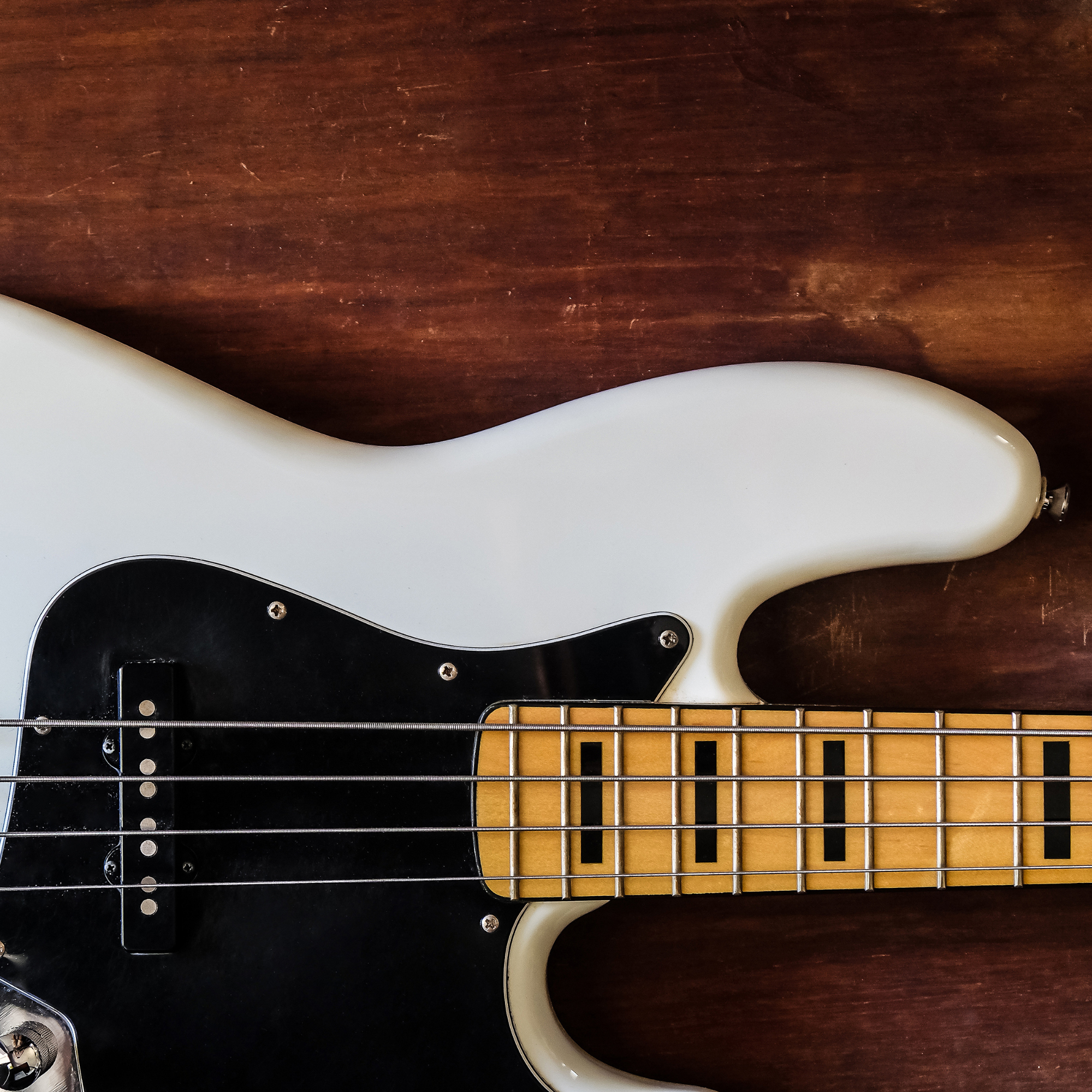 Close-up of a white electric bass guitar with black pickguard and maple neck, placed on a dark wooden surface.