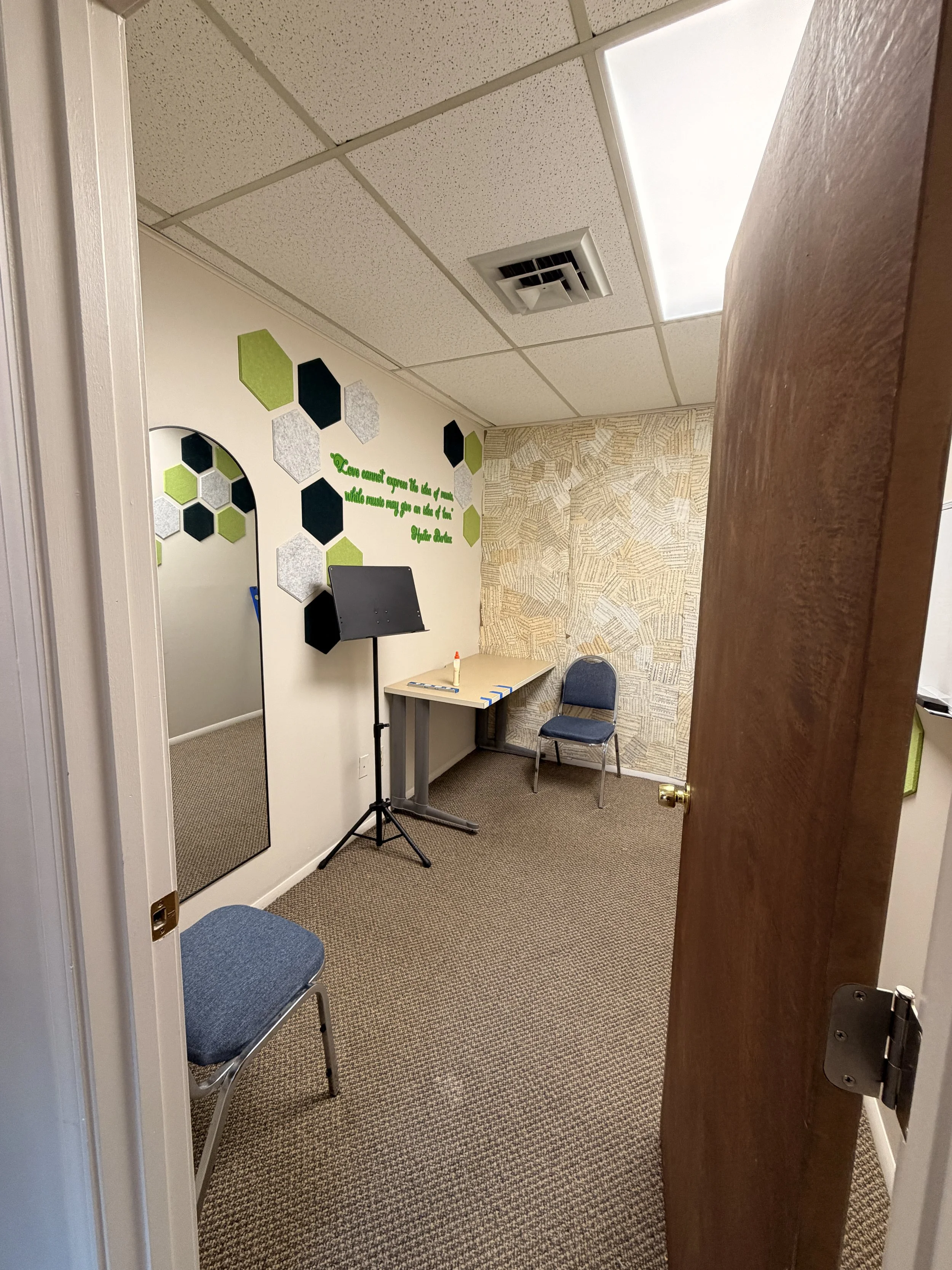 A small office or music practice room with beige walls, a mirrored wall, a small desk with a water bottle and some tape, two blue chairs, a music stand, wall decor with black, gray, and green hexagons, a quote painted in green, and a ceiling with a fluorescent light and an air vent.