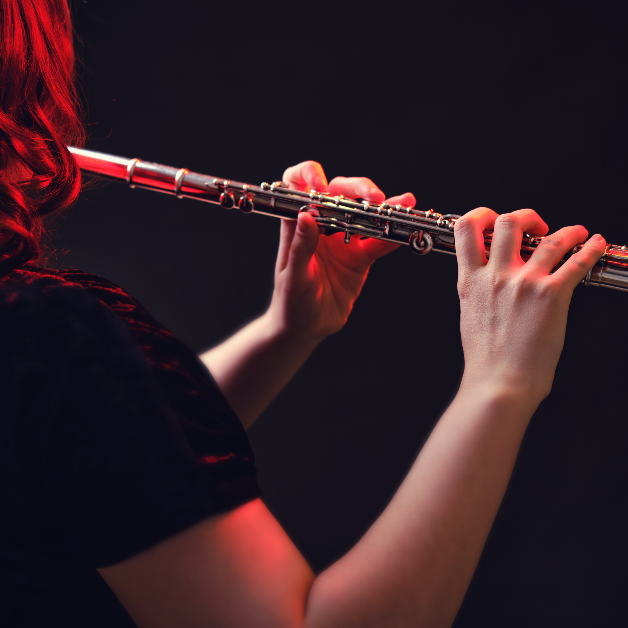 Person playing a silver flute in low light, highlighted by red lighting.