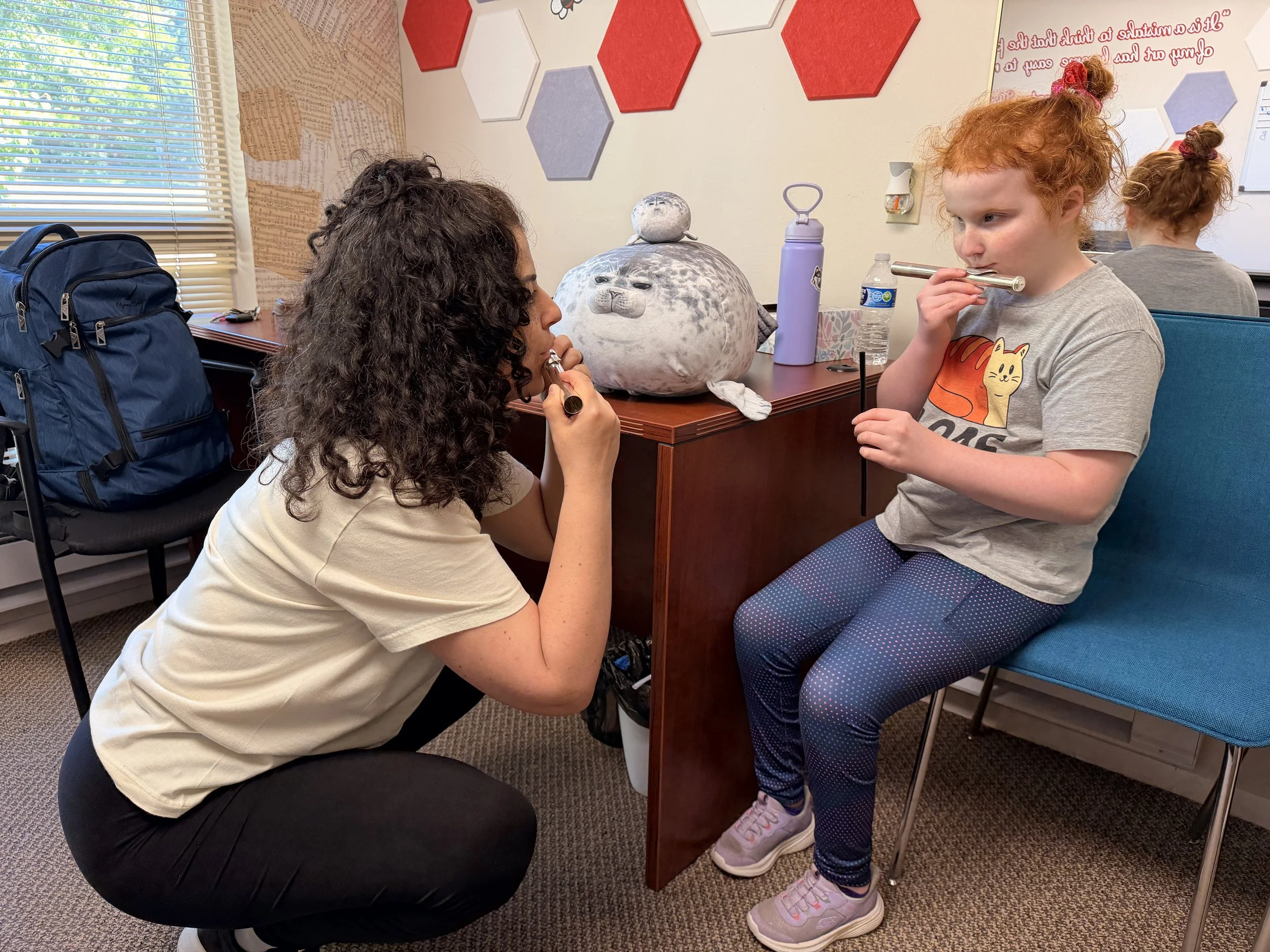 A woman kneeling and a girl sitting down playing recorders in a classroom. The girl is wearing a gray t-shirt with a tiger graphic, and the woman is wearing a cream shirt. The classroom has a window with blinds, colorful hexagonal wall decorations, and various objects on the desk including a plush toy, water bottles, and a backpack.