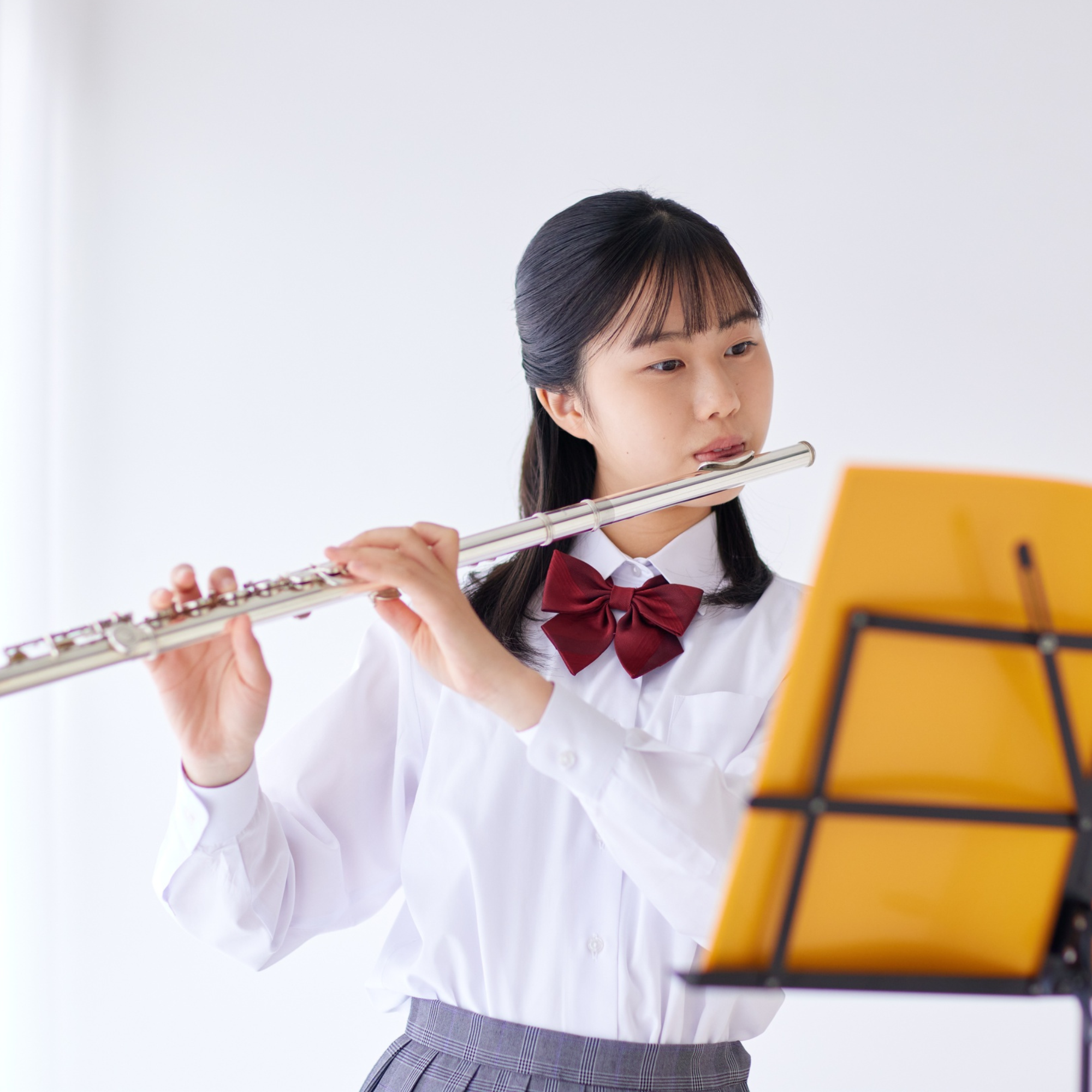 Young girl in school uniform playing a silver flute while reading sheet music on a yellow music stand.