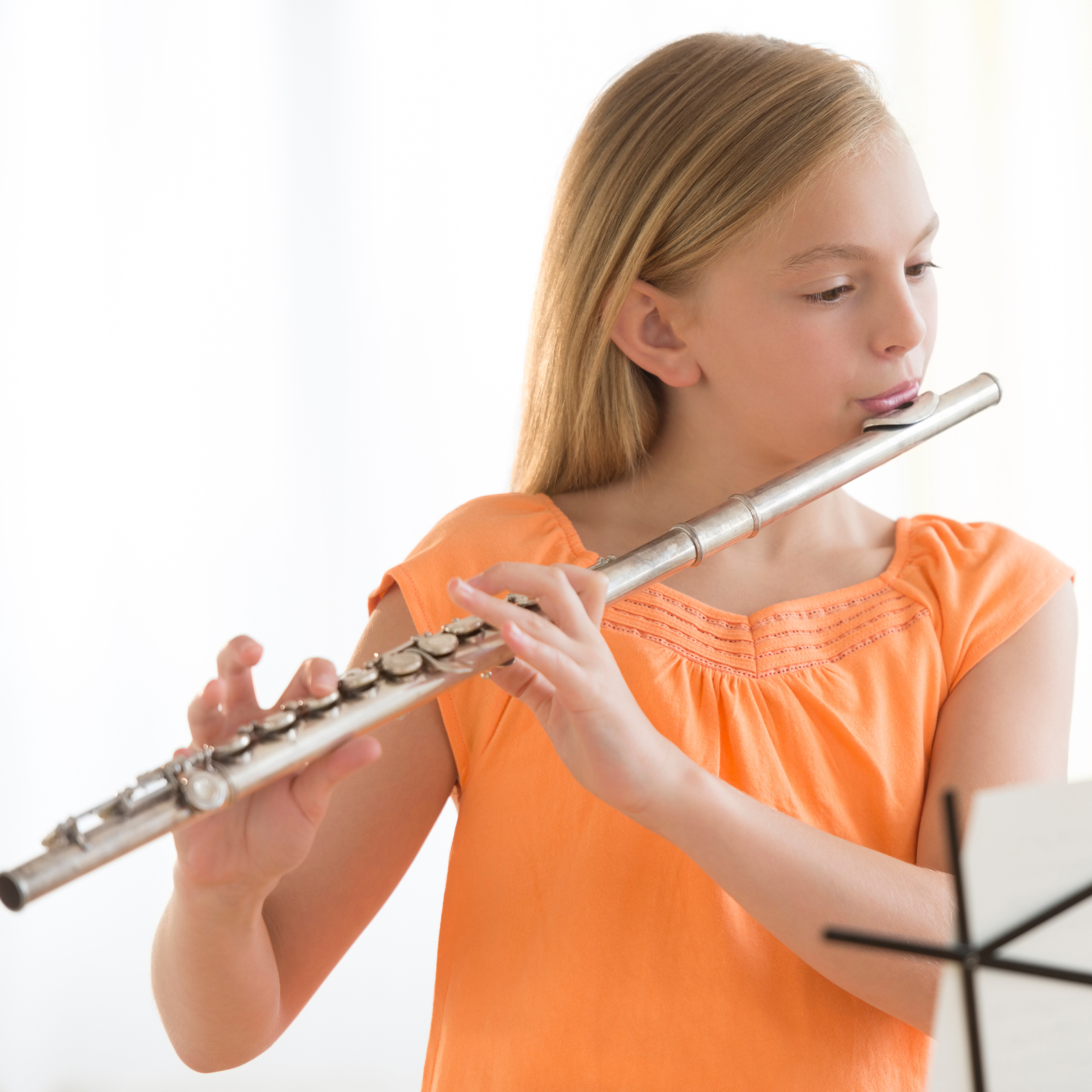 Young girl playing the flute while looking at sheet music, wearing an orange sleeveless top.