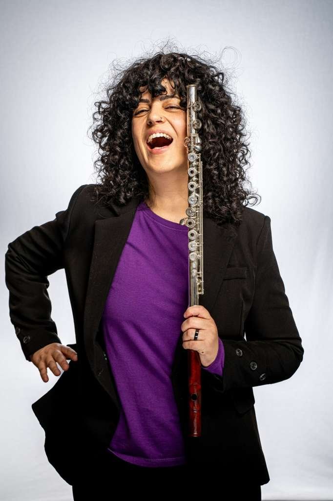 A woman with curly dark hair, wearing a purple shirt and black blazer, holding a flute vertically in front of her face, with a joyful expression.