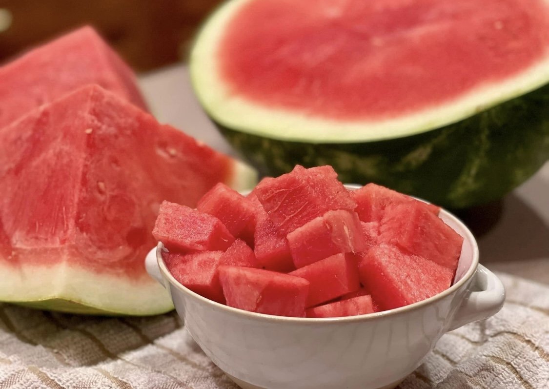 Pieces of watermelon in a small white bowl, with a sliced open watermelon and a watermelon wedge in the background.