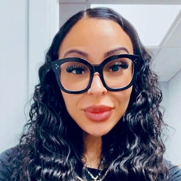 A woman with long, dark hair, wearing large, rhinestone-studded glasses, leopard print jacket, black top, and layered gold necklaces, posing against a light blue background.