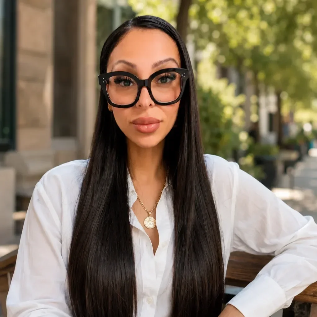 A woman with long, dark hair, wearing large, rhinestone-studded glasses, leopard print jacket, black top, and layered gold necklaces, posing against a light blue background.