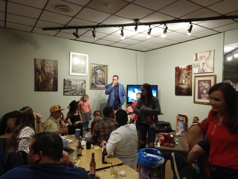 People gathered in a room, some sitting at a table with food and drinks, engaging with two presenters standing near a television. The room has framed artwork on the walls, overhead track lighting, and a lively atmosphere.