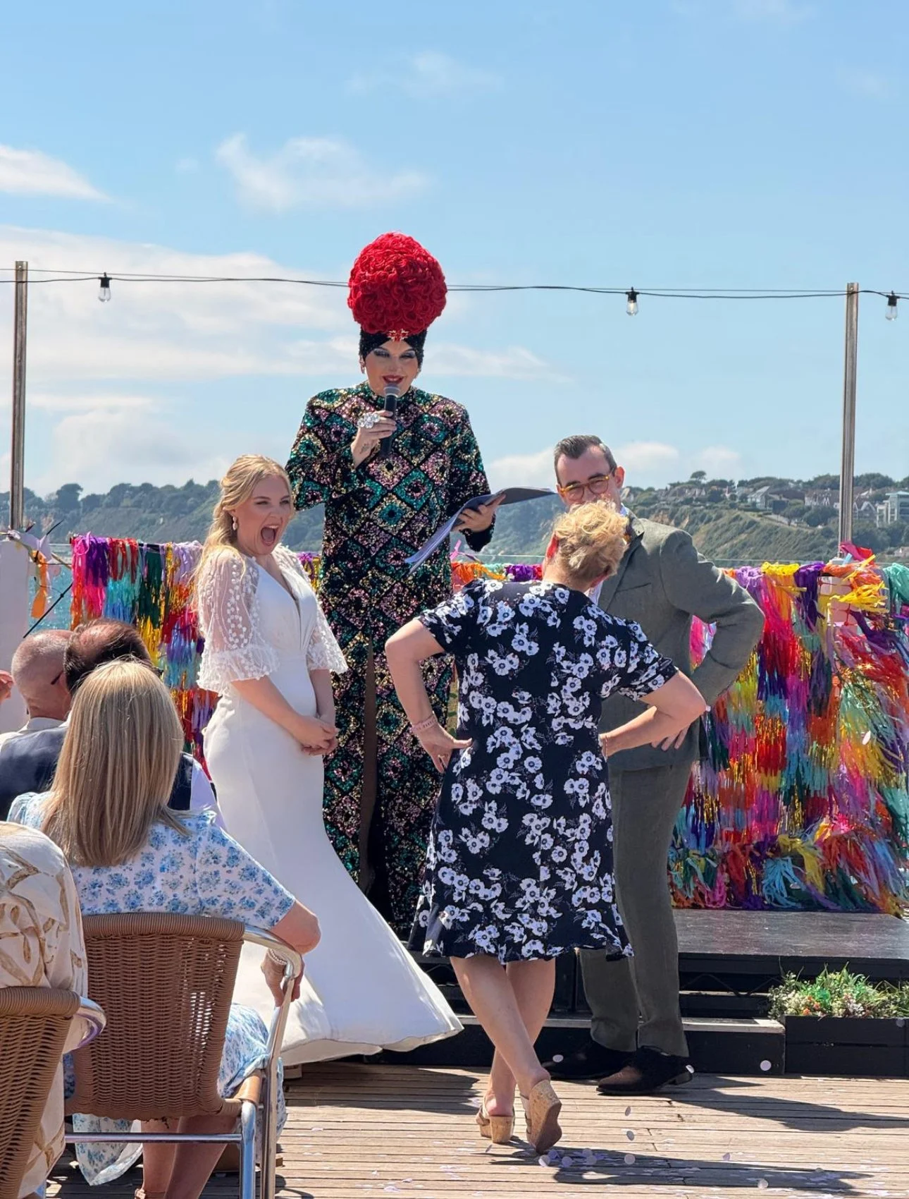 A drag queen in an ornate dress with a tall red hairpiece holding a microphone at an outdoor event, surrounded by guests seated and standing on a wooden deck with colorful decorations and a scenic hillside in the background.