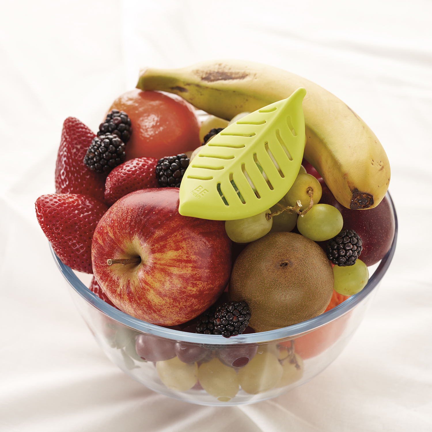 A glass bowl filled with various fruits including apples, bananas, strawberries, blackberries, green grapes, and kiwi, with a green pleated fruit peeler resting on top.