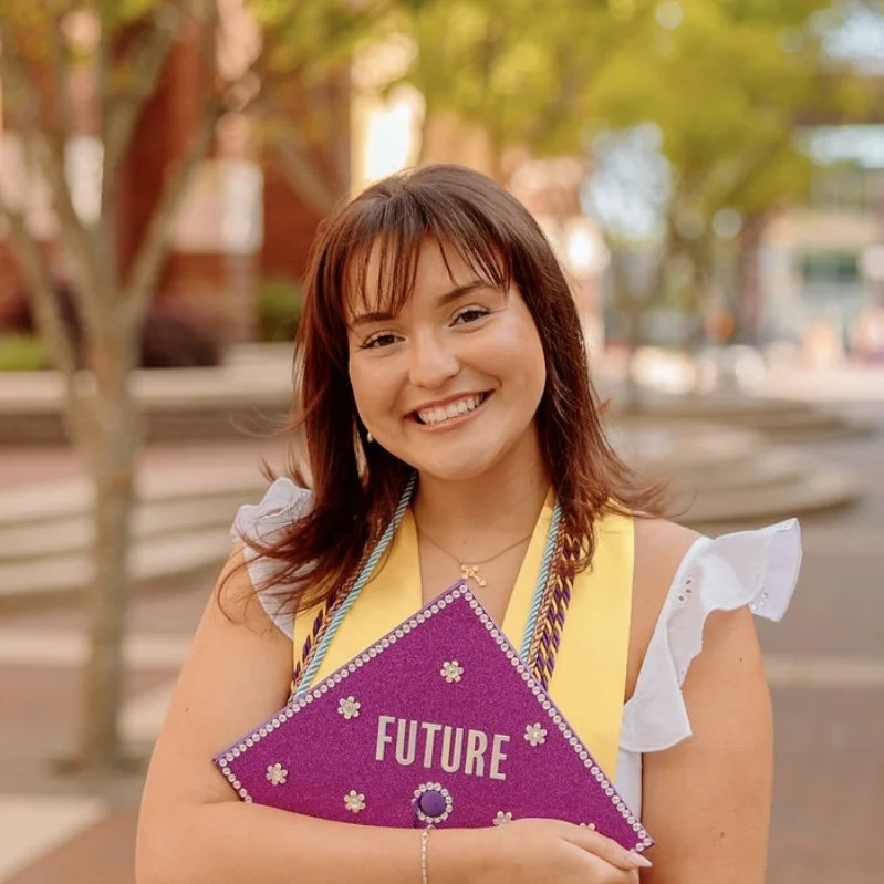 A young woman smiling and holding a decorated graduation cap with the word 'Future' written on it, outdoors with trees and buildings in the background.