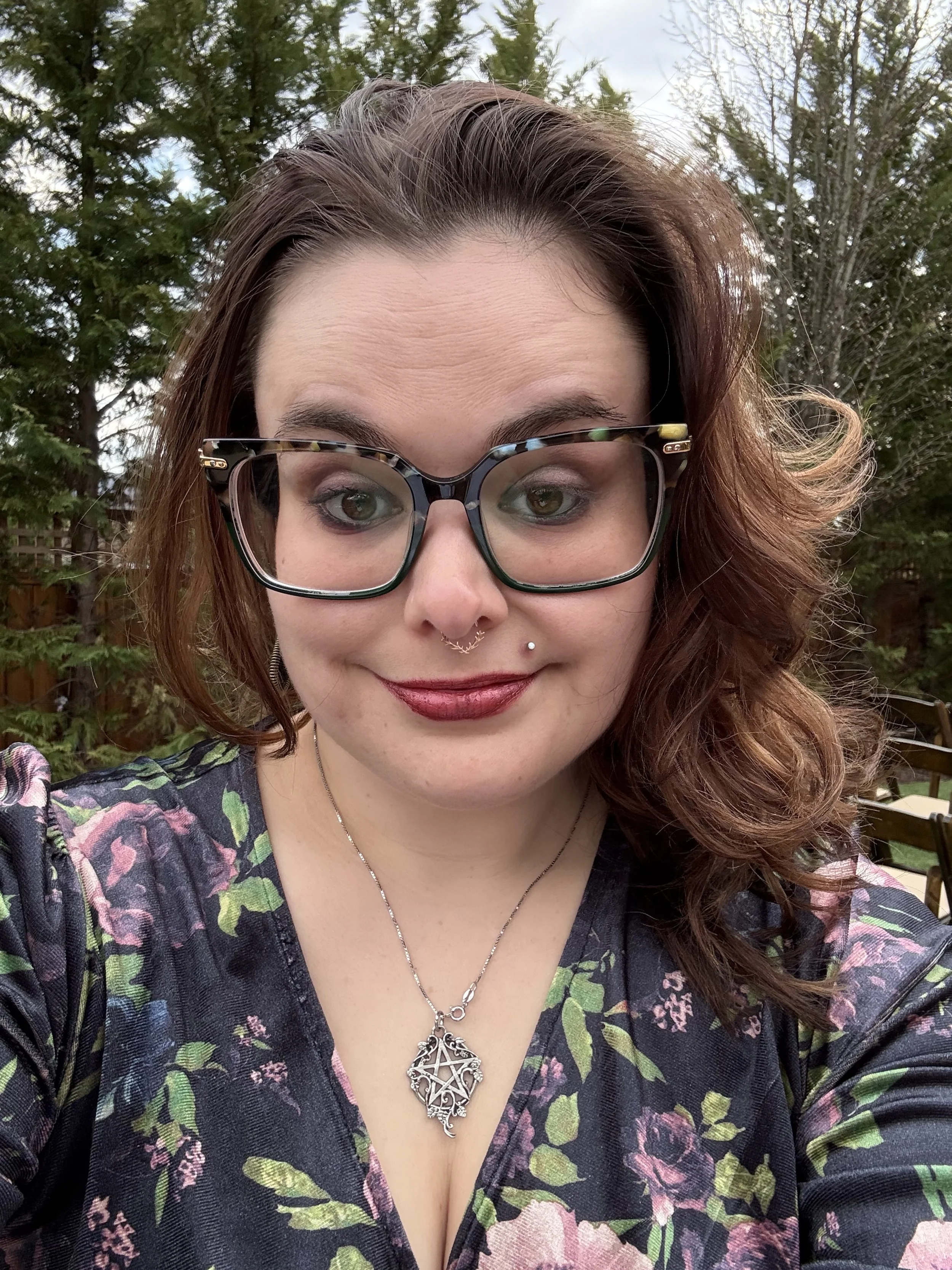 A woman with wavy brown hair, wearing large black glasses, a dark floral top, and a necklace with a decorative silver pendant, taking a selfie outdoors with green trees in the background.
