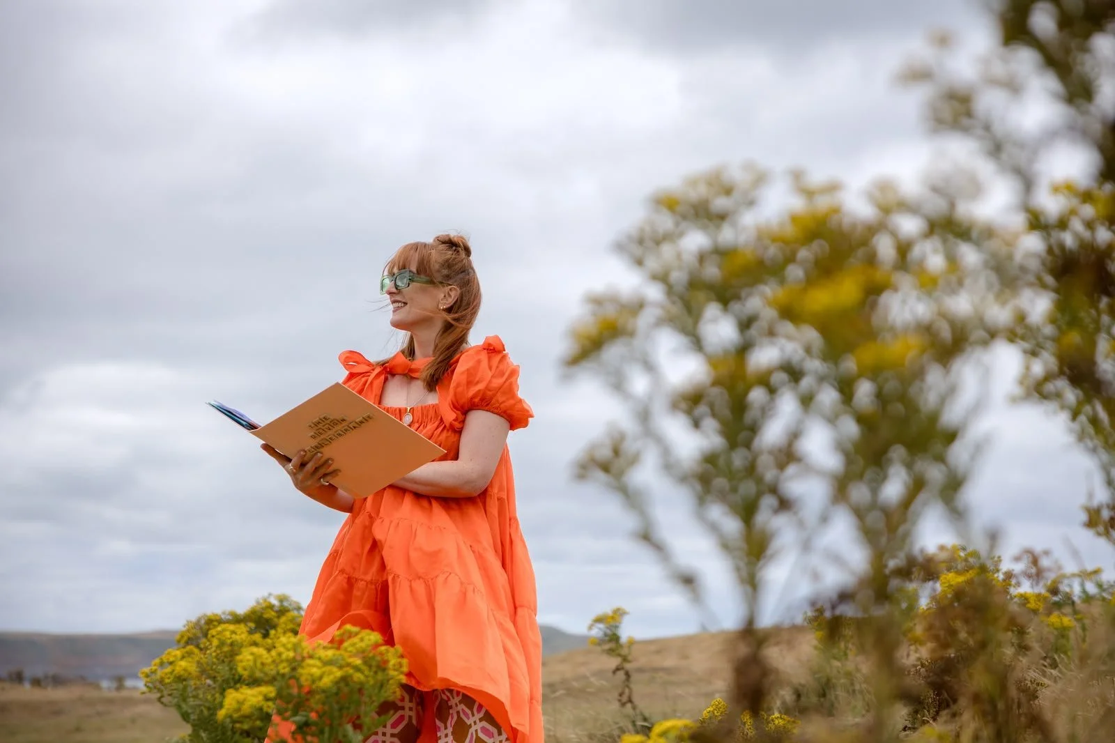 Smiling woman with red hair and sunglasses, wearing an orange dress, holding a book outdoors in a field with yellow flowers and a cloudy sky.