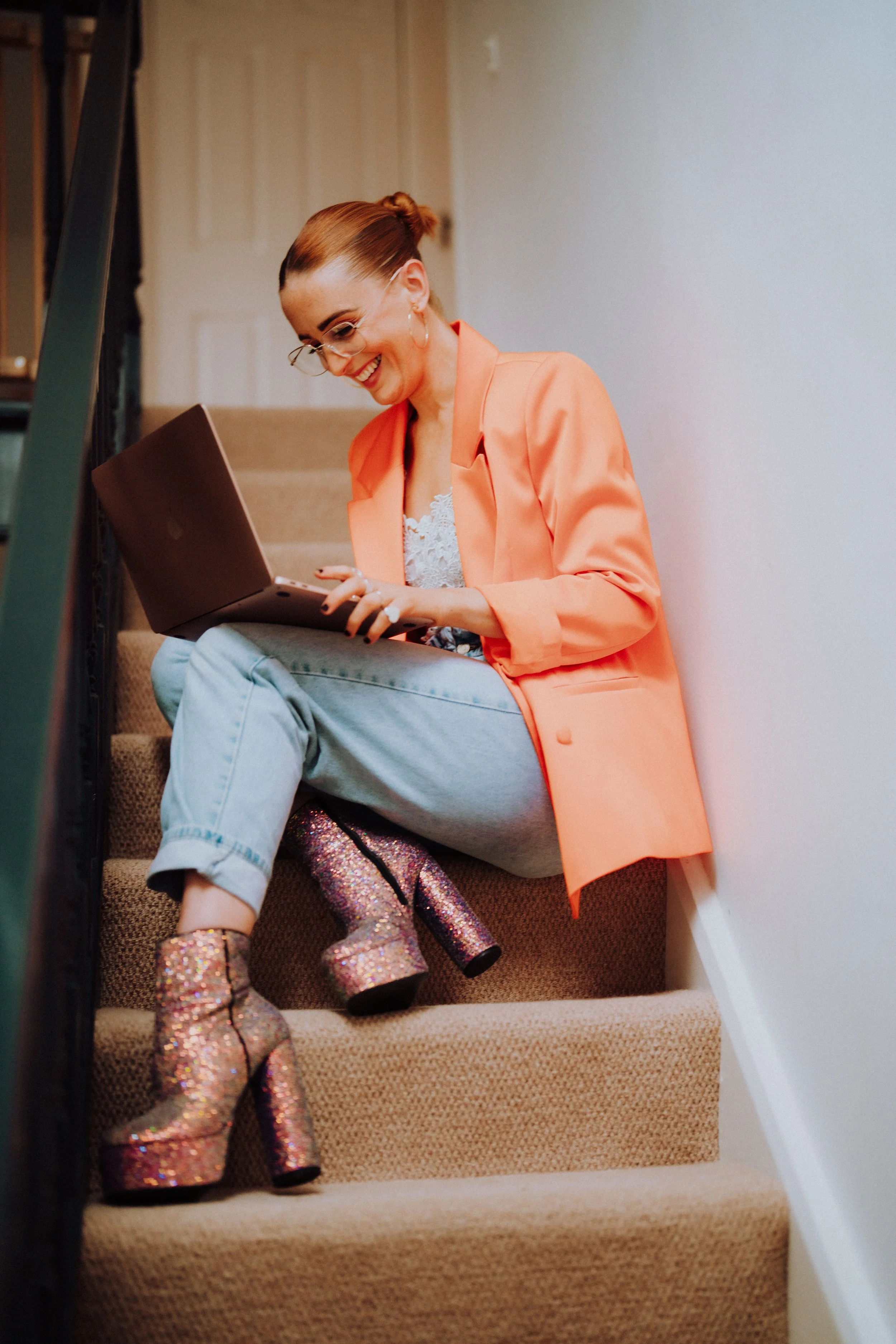 A woman with glasses and a bright orange blazer sitting on carpeted stairs, using a laptop, with glittery high-heeled boots.