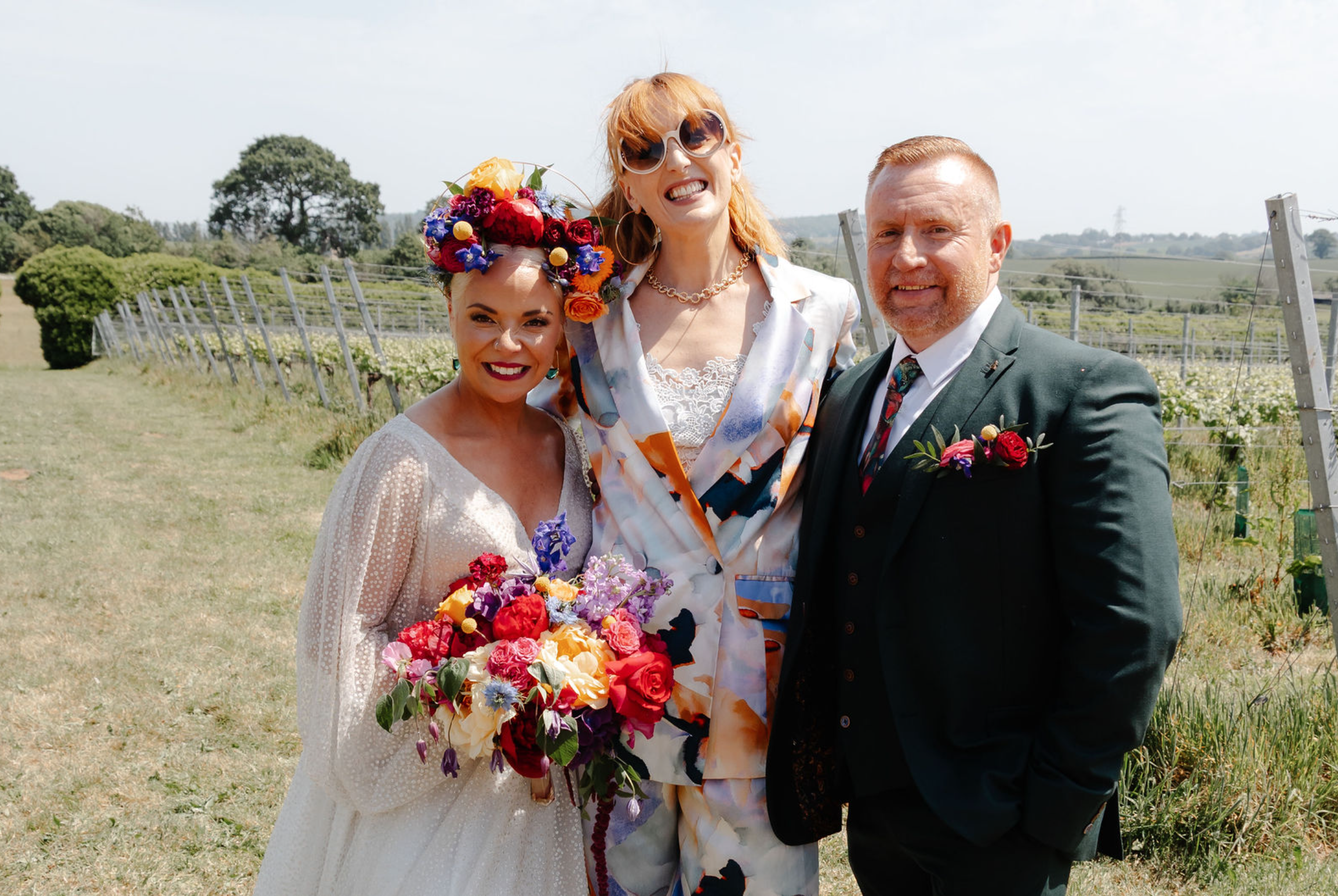 A bride and groom embracing during an outdoor wedding ceremony, with guests in the background and a house with trees and bushes in the distance.