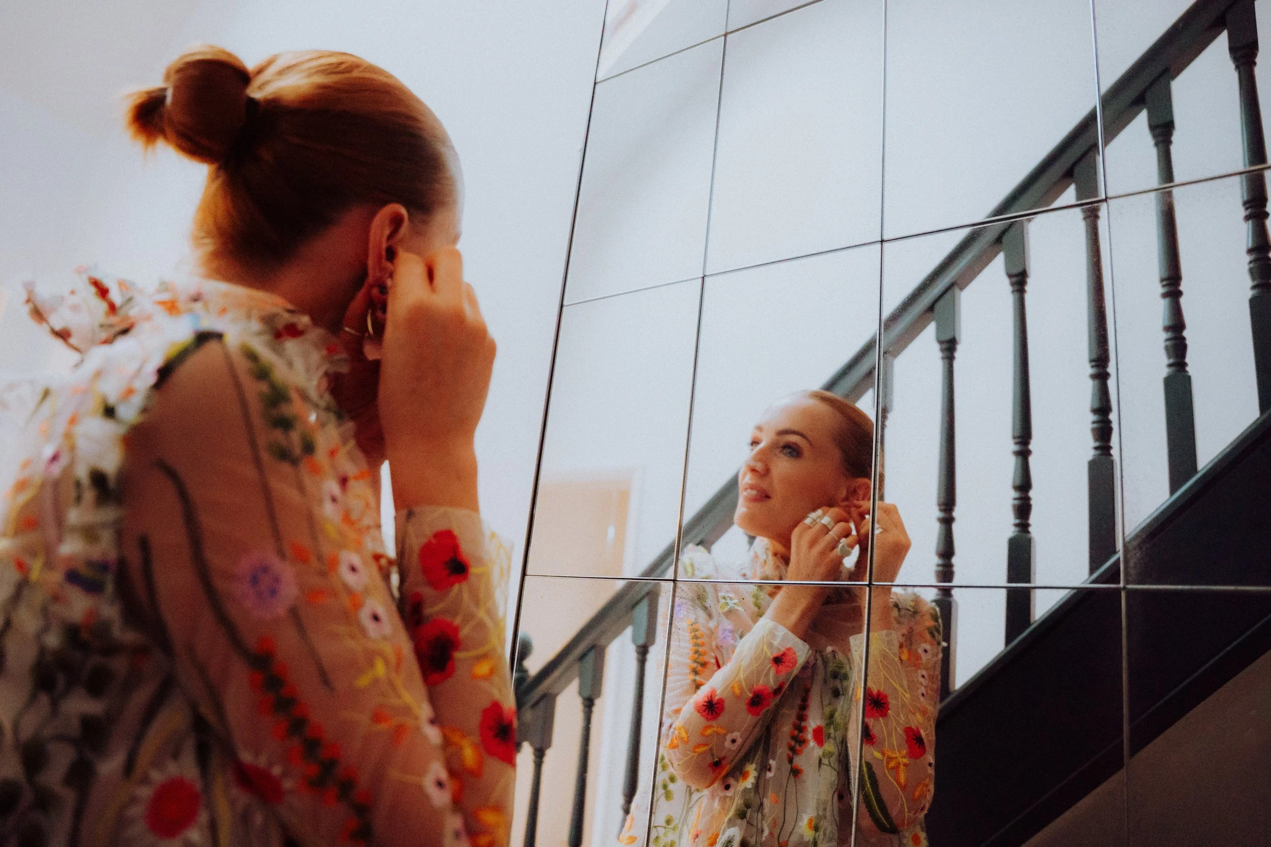 A woman with red hair wearing a sheer, floral embroidered top, standing in front of a mirror on a staircase, adjusting her earrings.