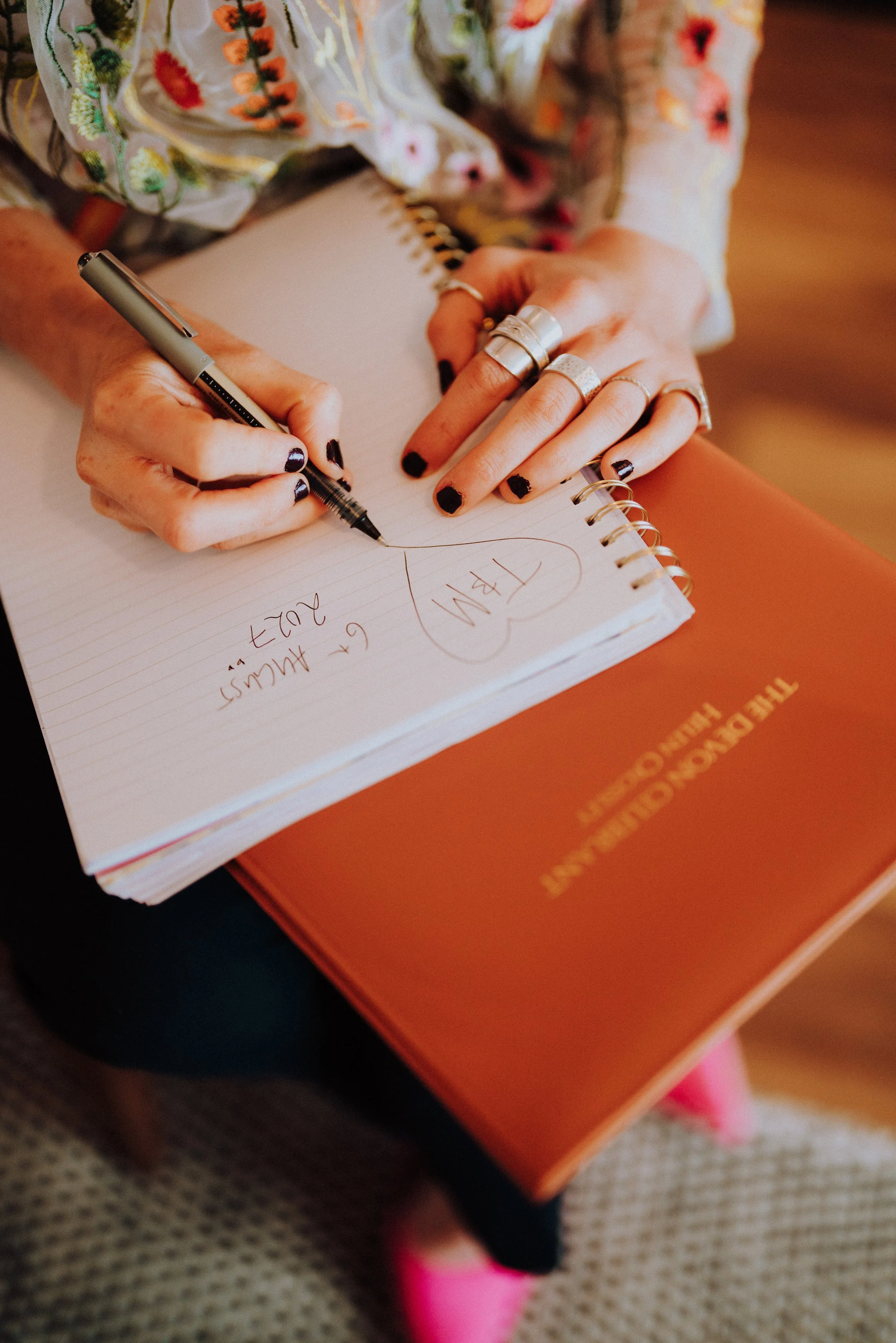 A person with dark nail polish and numerous rings is writing in a spiral notebook with a black pen. The notebook is on top of a bright orange book titled 'The Courage to Be Disliked' by Ichiro Kishimi and Fumitake Koga. The person is sitting at a table, and part of their pink shoe is visible.
