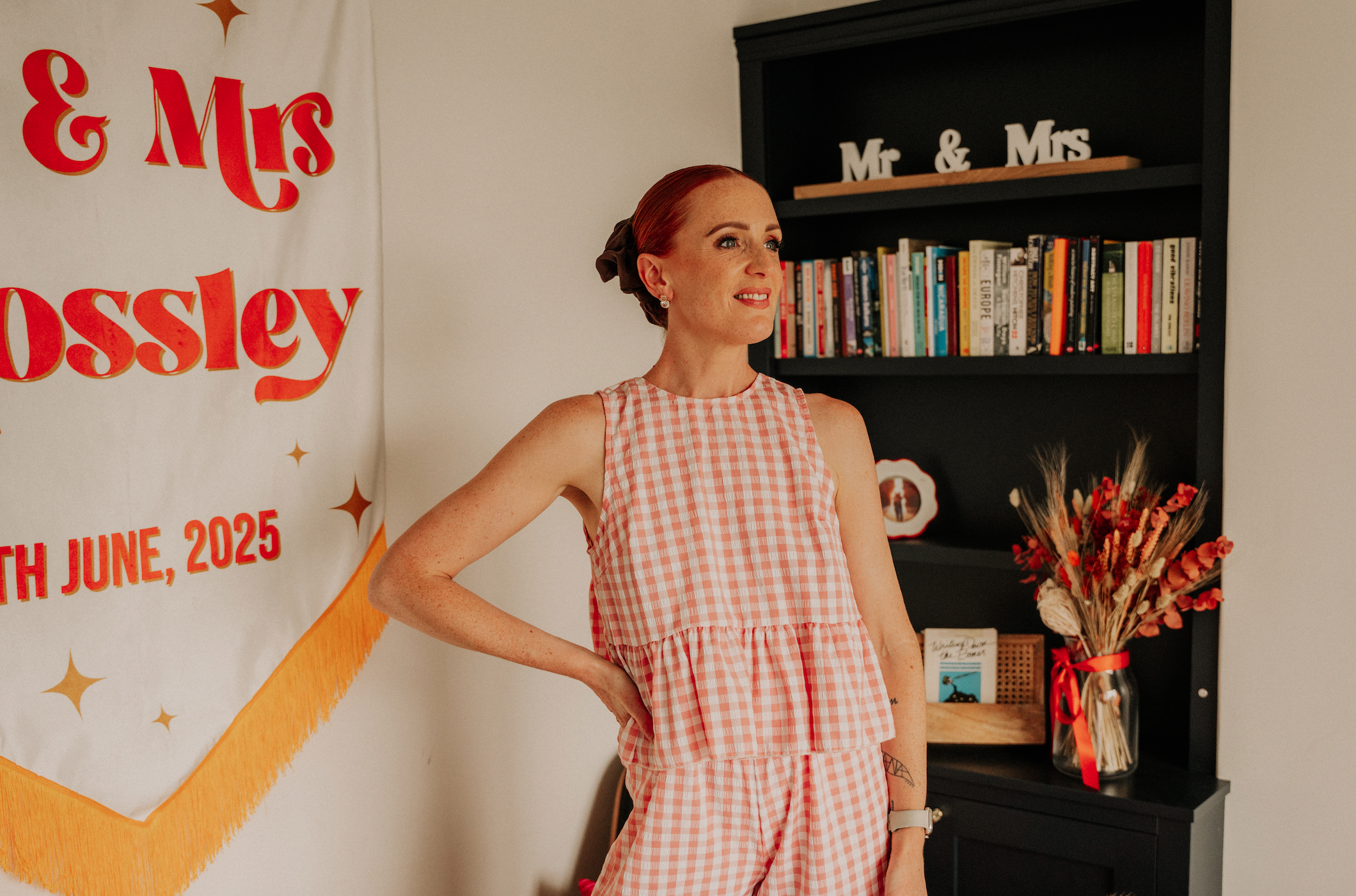 Woman with red hair in a bun wearing a sleeveless pink and white checkered dress, standing indoors next to a black bookshelf with books and decorative items, with a wedding banner that reads '& Mrs Ossley' and 'June, 2025' in the background.
