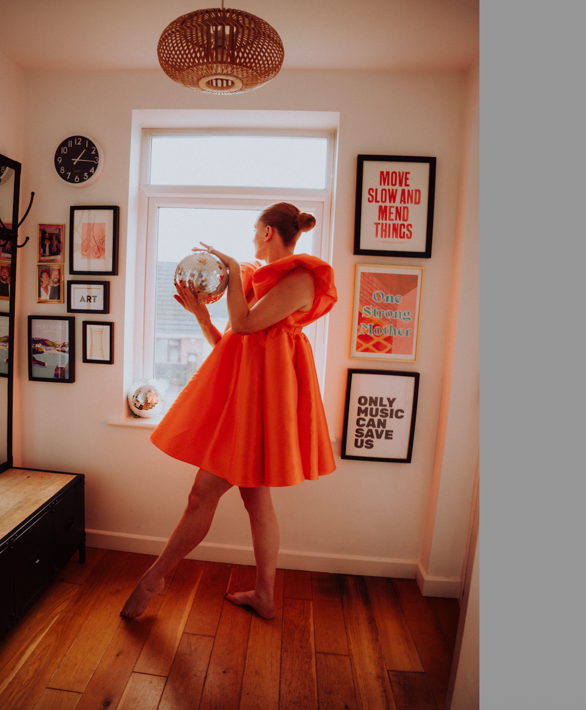 A woman in an orange dress holding a disco ball, standing barefoot on a wooden floor near a window in a decorated room with framed art and quote posters.