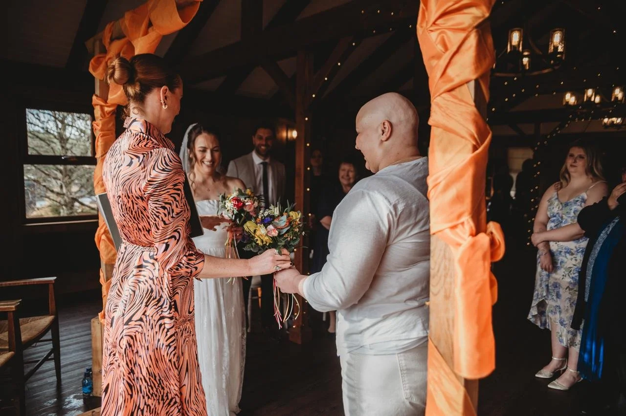 A woman with red hair wearing a sheer, floral embroidered top, standing in front of a mirror on a staircase, adjusting her earrings.