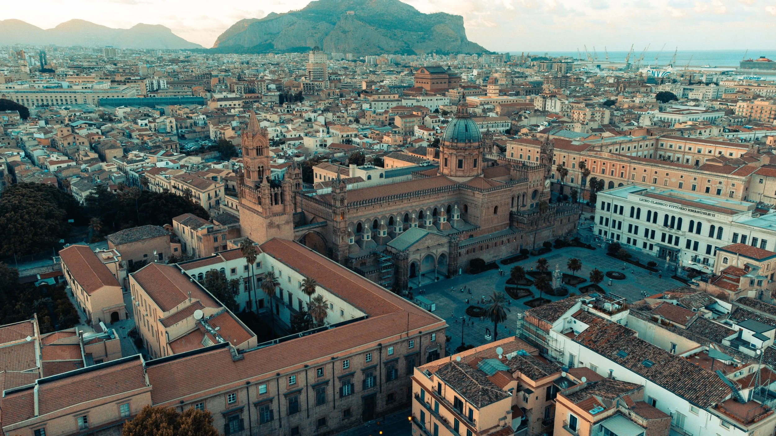 Luftaufnahme einer Stadt mit historischem Dom und umliegenden Gebäuden, Berge im Hintergrund, Küstenlinie mit Hafenanlagen im Abstand.