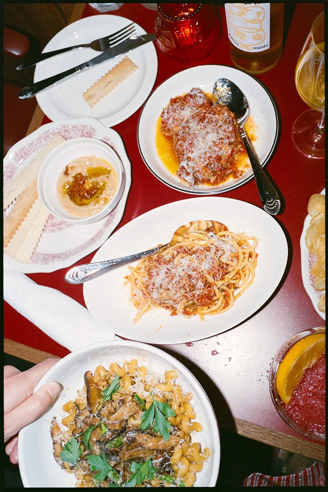 Italian meal with spaghetti, chicken parmesan, a bowl of beans with herbs, and drinks including beer, wine, and a cocktail with lemon wedge on a red table.